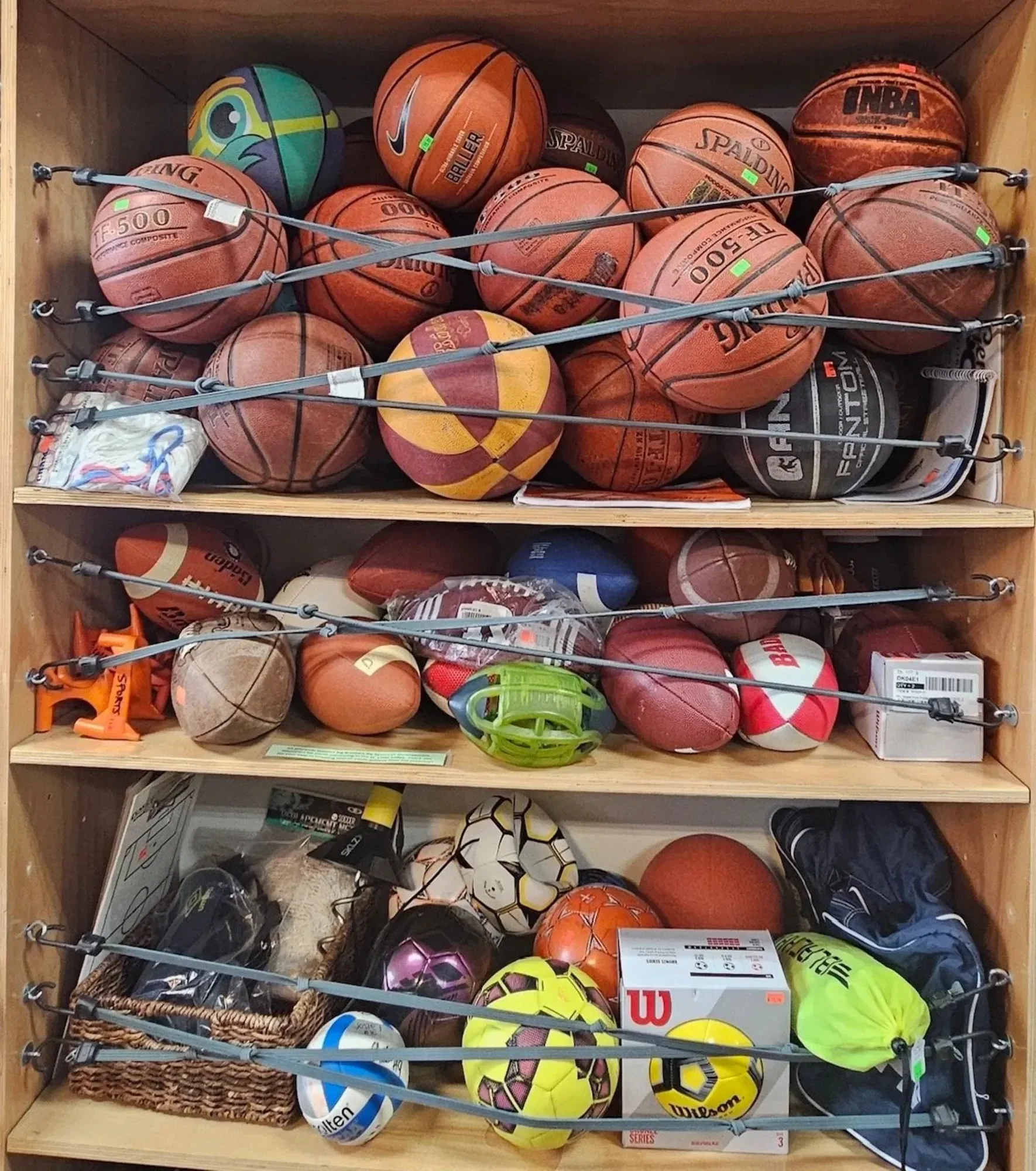A wooden shelf filled with various basketballs and soccer balls, some in baskets or bags, with elastic straps securing them.