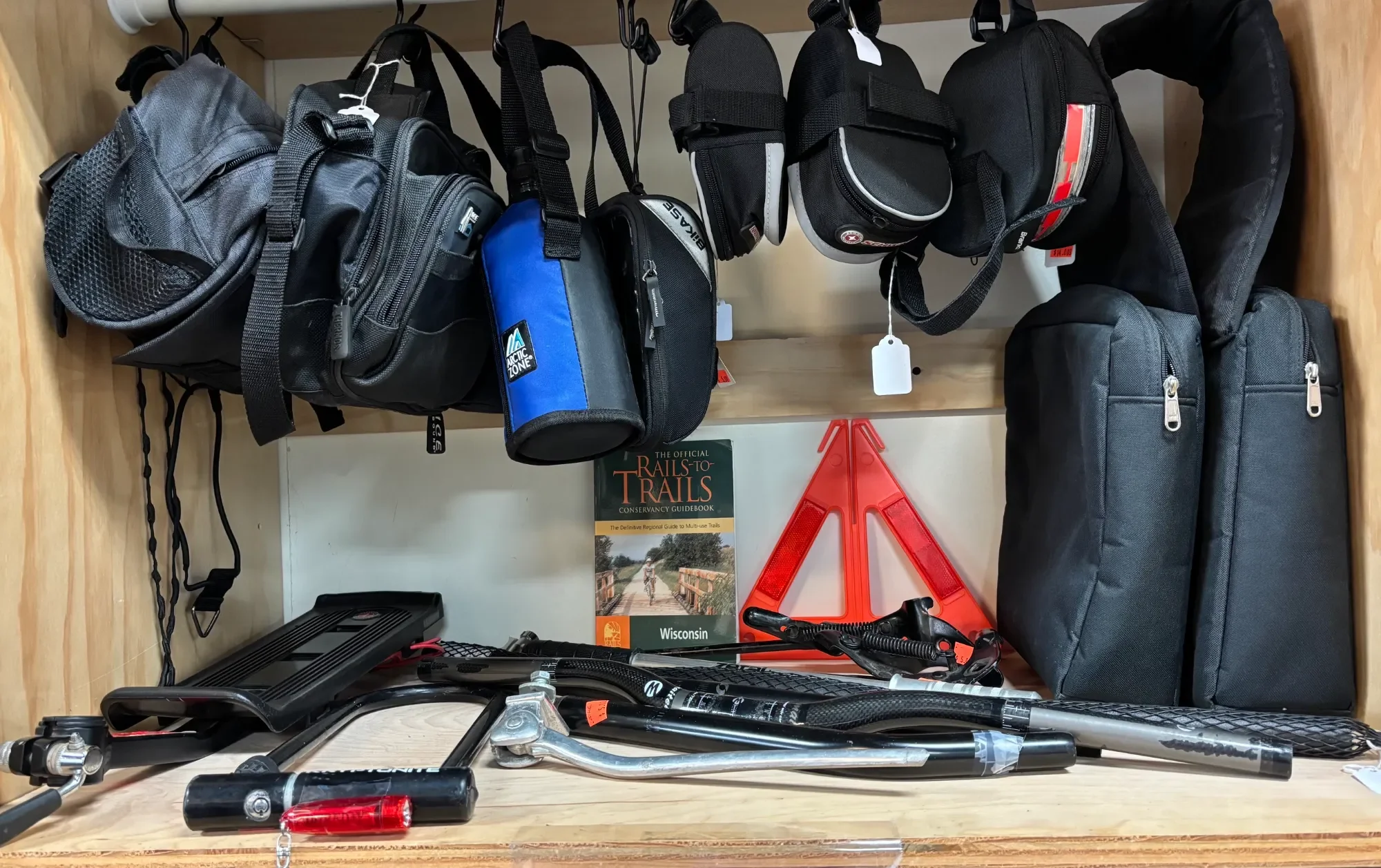 A wooden shelf holding various items including backpacks, a book titled 'Trails to Trails', a red safety triangle, a bike helmet, and a piece of bike equipment, with a white wall in the background.