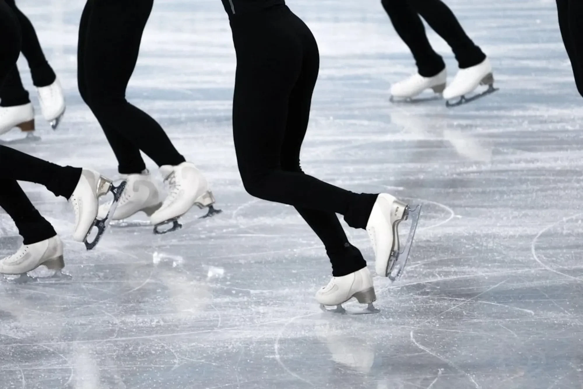 Close-up of multiple figure skaters' legs and feet wearing white ice skates on an ice rink, captured during a skating practice or performance.