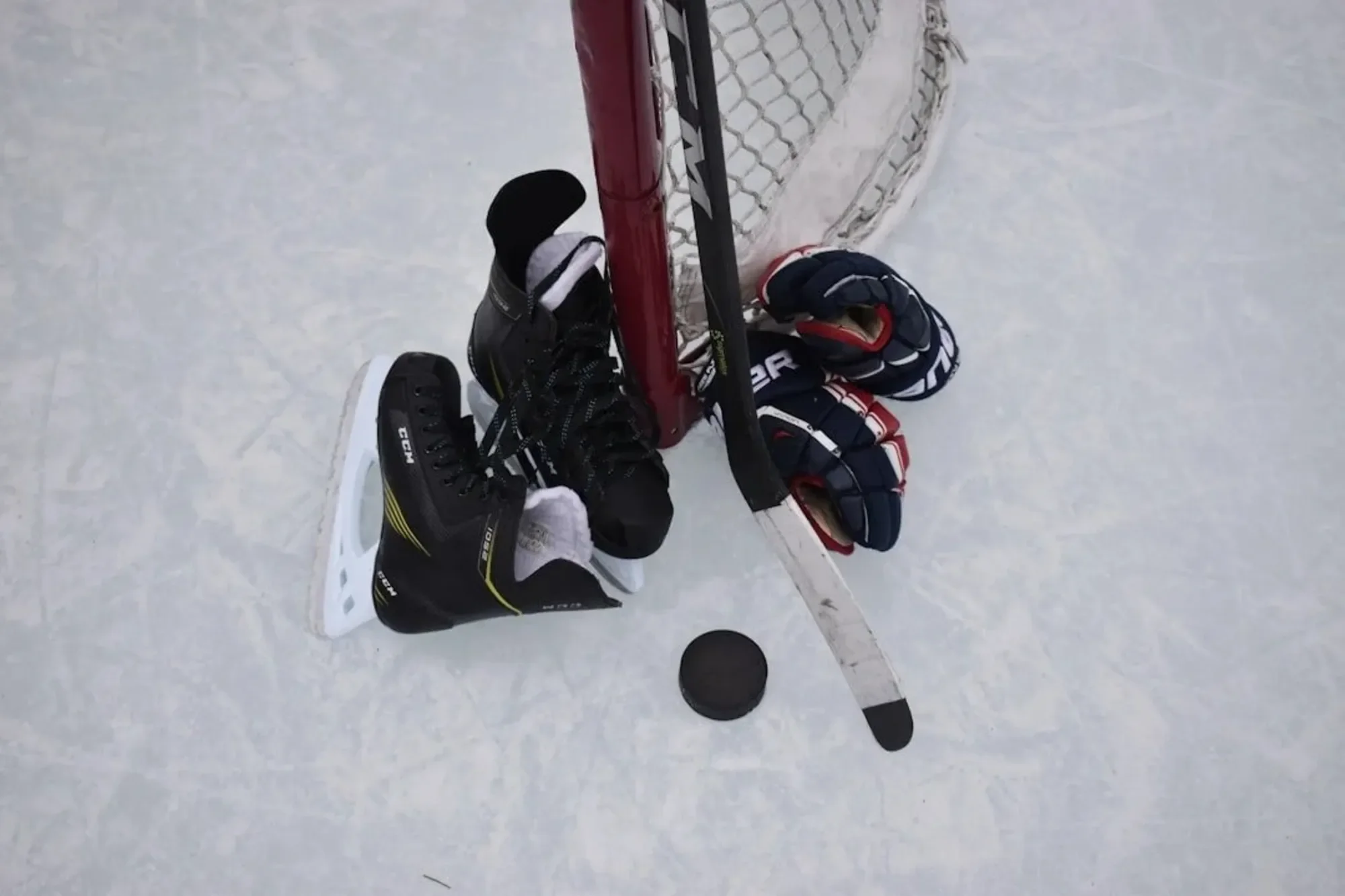 A hockey game scene viewed from above showing four hockey players, two on each side of the goal, with sticks and puck on the ice. One player is in black gear skating near the goal, while the other players are in different uniforms. The hockey net with red posts and white netting is visible.