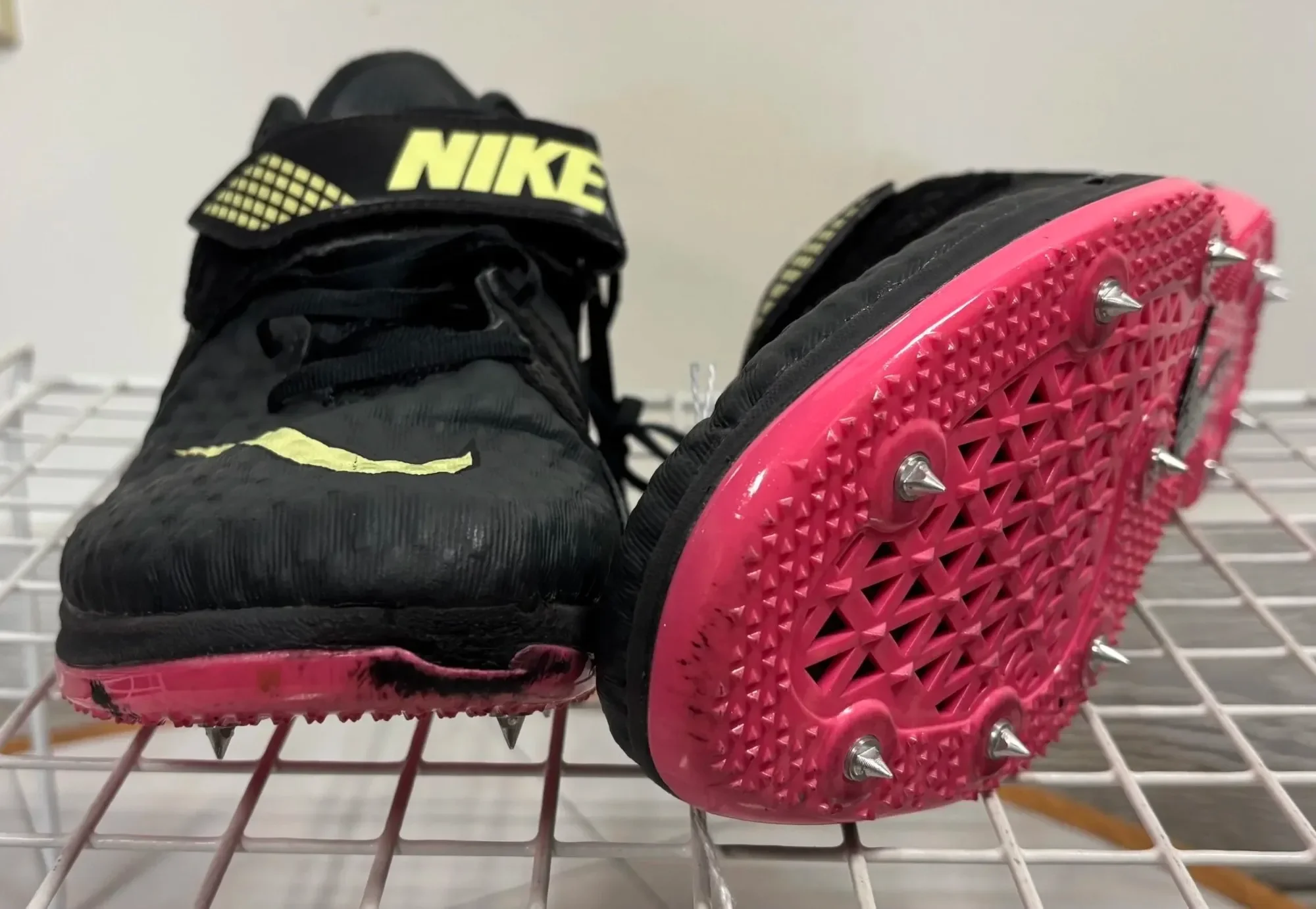 A pair of black Nike sneakers placed on pink spiked roller skate wheels with metal spikes on a white wire rack.