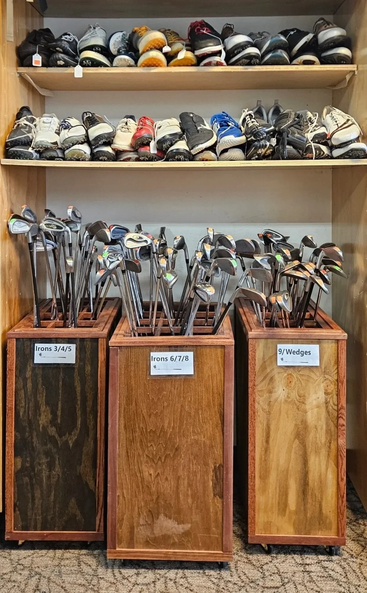 Golf shoes on the top shelf, organized golf clubs in wooden bins labeled for different club types in front, set on a carpeted floor.