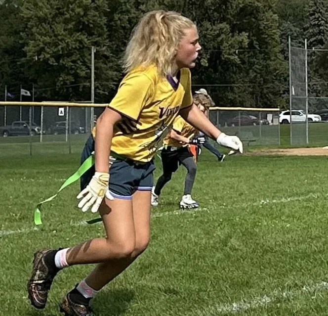 A young girl wearing a yellow sports jersey, shorts, and soccer cleats, with gloves on, is running on a grassy field during a soccer game.