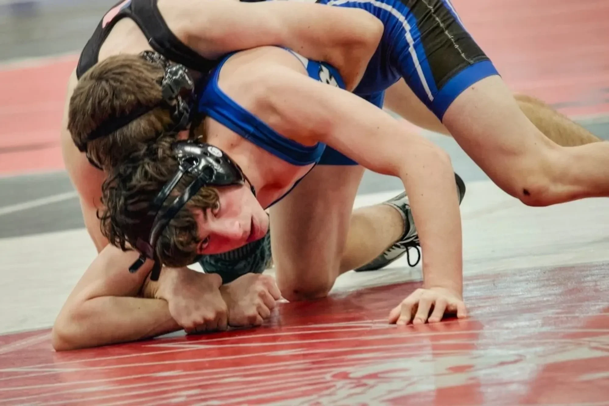 Two wrestlers, one wearing a black singlet and the other a blue singlet, engaged in a wrestling match on a red and gray mat. Both are wearing protective headgear, and the wrestler in blue is on top, pinning the other to the mat.