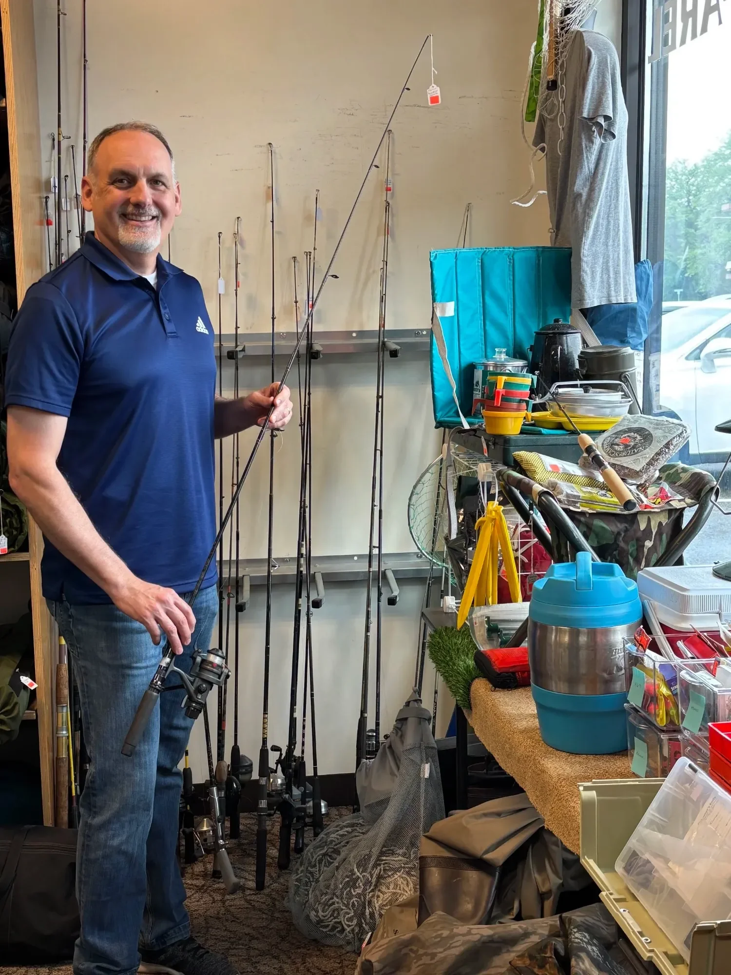 A man smiling in a fishing gear store, holding a fishing rod, with fishing rods displayed on the wall behind him, and various fishing and outdoor gear scattered across a counter and shelves.