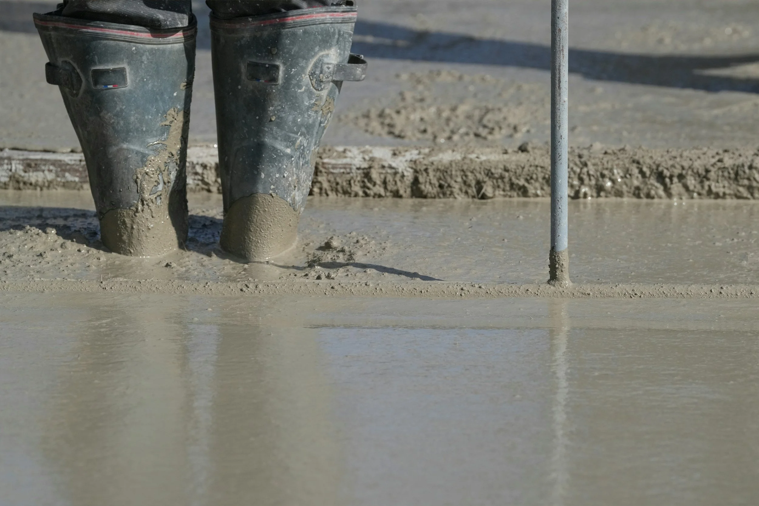 Close-up of a concrete workers legs wearing rubber boots standing on wet concrete near a metal rod at a construction site.