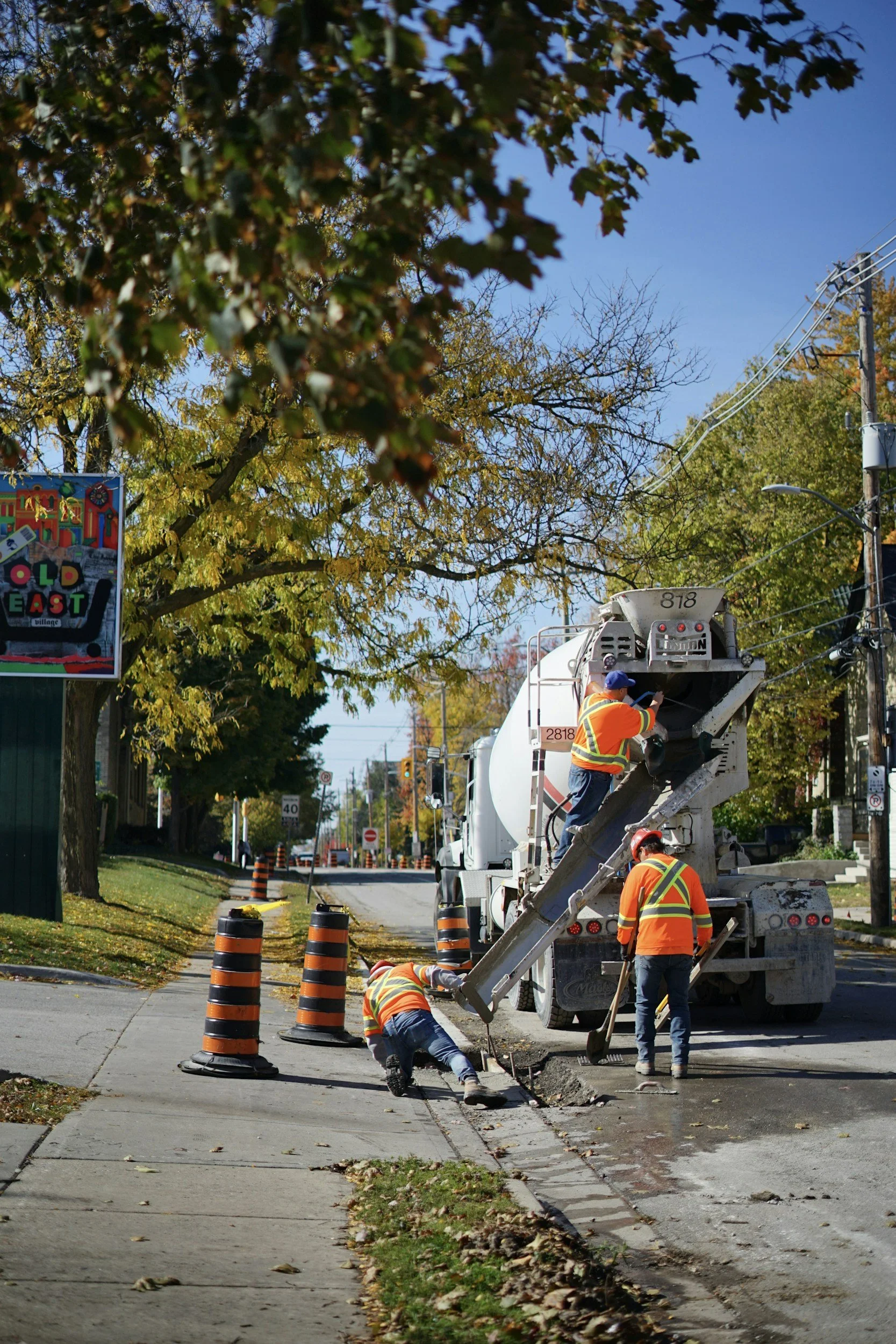 Workers in orange safety vests and helmets repairing a section of sidewalk with a truck and construction cones around the work area. Trees with fall foliage line the street.