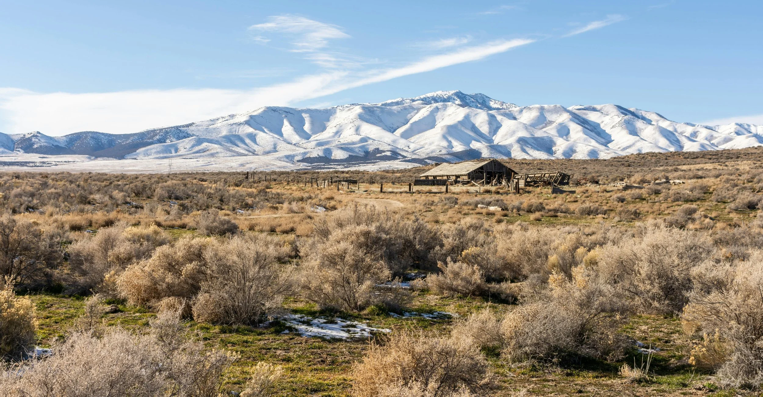 A wide landscape view of a desert with sparse bushes in the foreground, a dilapidated barn in the middle ground, and snow-capped mountains in the background under a blue sky with wispy clouds in Rush Valley, Utah.