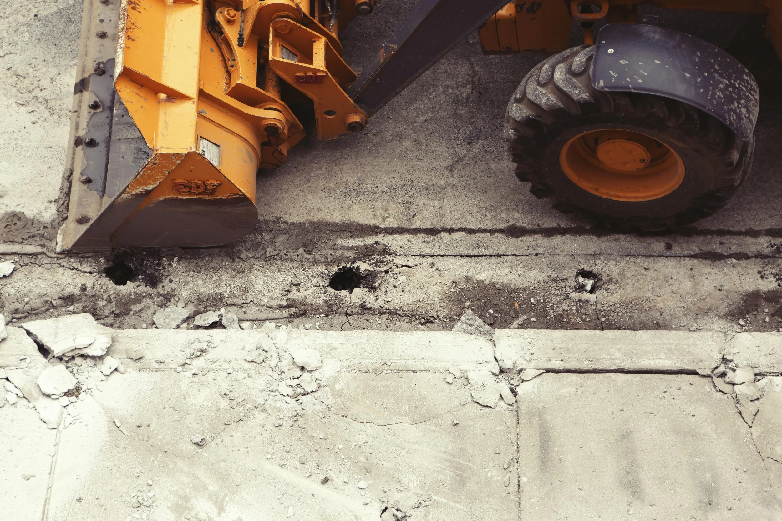 Close-up of a construction site with a yellow heavy-duty machine on a concrete surface, with cracks and debris around.