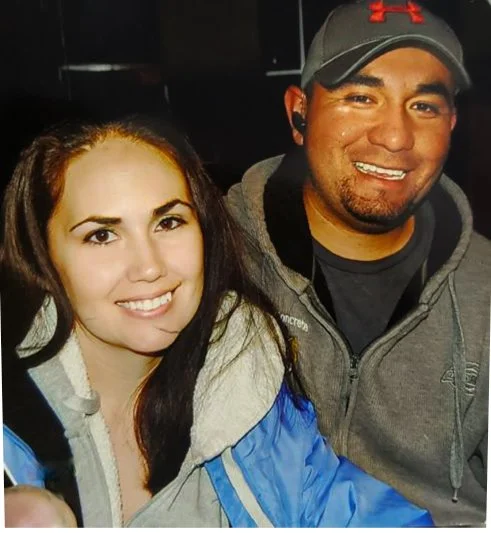 Nicole and Daniel Ramirez-picture of the owners of Ramirez Concrete and Design, LLC. Two people smiling, a woman with long dark hair in a light jacket and a man with a gray cap and hoodie, indoors with a dark background.