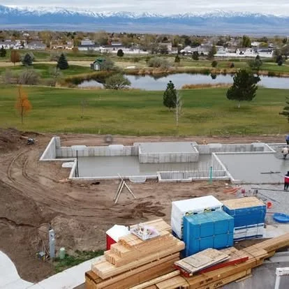 Construction site with a concrete foundation, wooden planks, and construction materials in the foreground. In the background, there is a grassy area with trees, small water bodies, and houses under a cloudy sky.