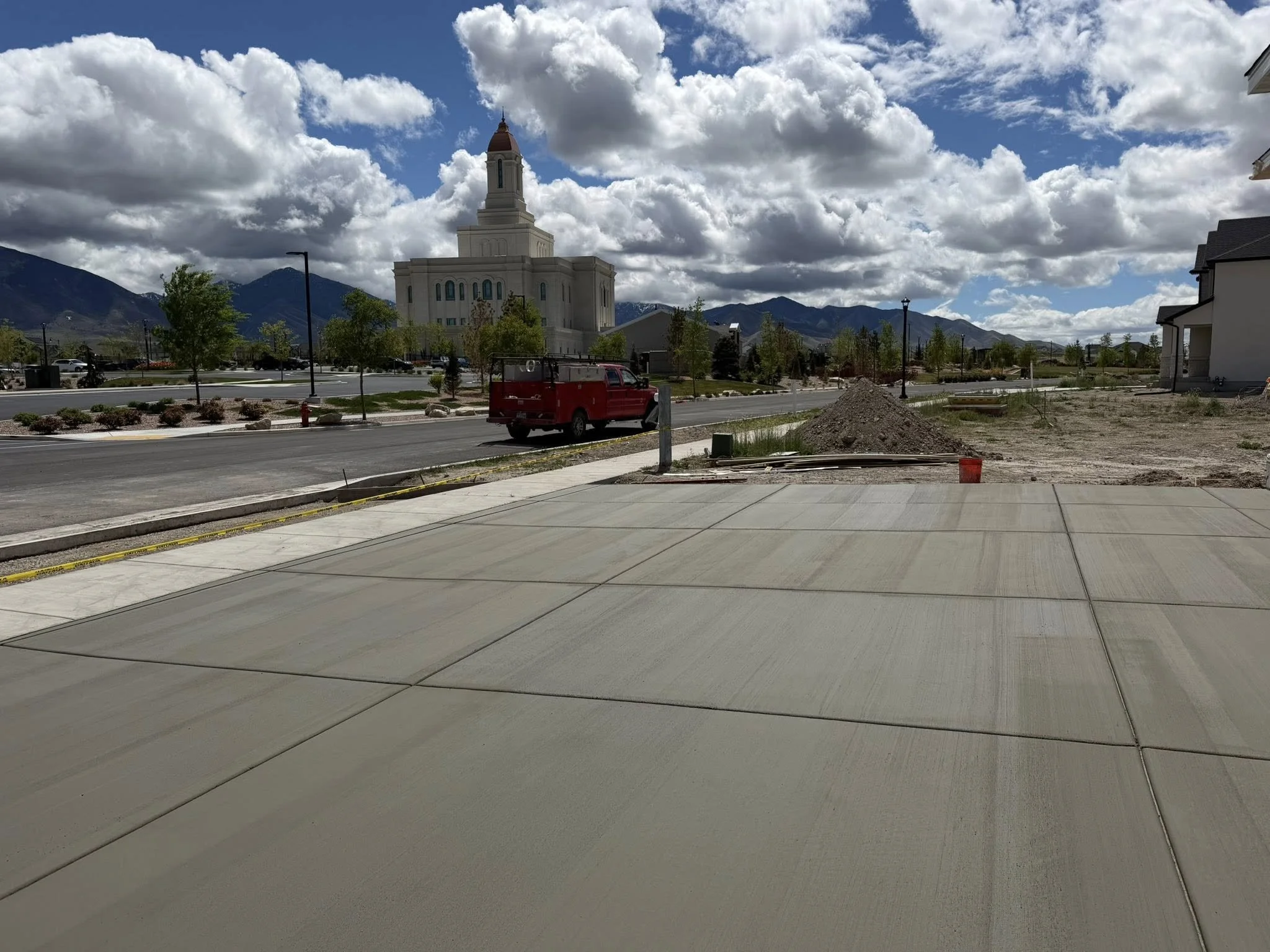 A large church with a tall steeple against a backdrop of mountains and cloudy sky. In the foreground, there is a paved area, small trees, a red utility truck, and a freshly poured concrete driveway.