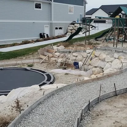 Concrete walkway preparation with curved forms, gravel base, and landscape rock features ready for installation.