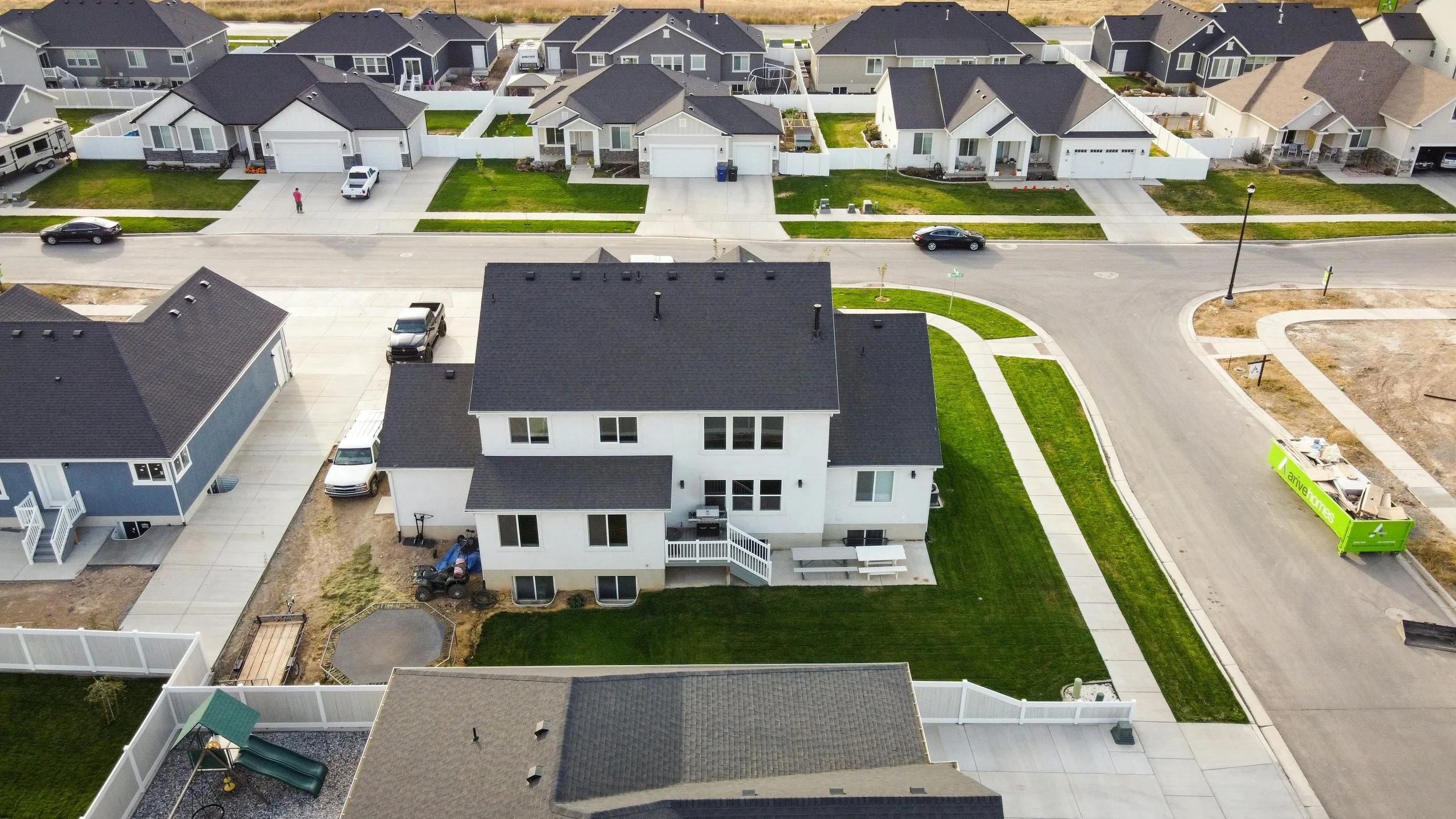 An aerial view of a suburban neighborhood with several modern houses, green lawns, parked cars, fences, and a curved street. Some houses are under construction or have construction materials visible.