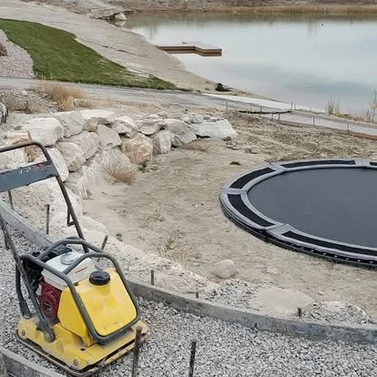 Concrete patio preparation near the water with compacted base, curved forms, and equipment ready for installation.