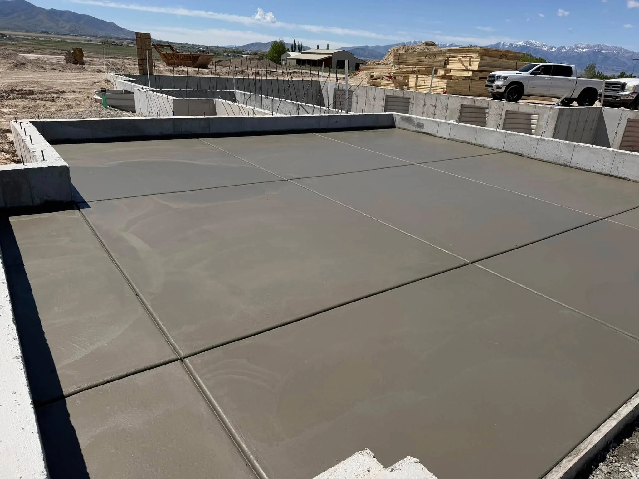 A construction site with freshly poured concrete foundation, surrounded by construction materials and trucks, with mountain scenery in the background.