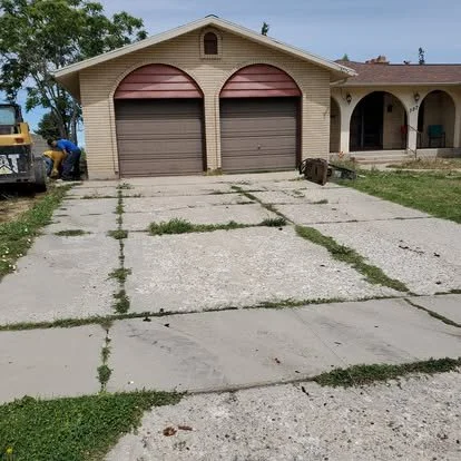 A driveway with overgrown weeds between concrete slabs in front of a suburban house with a two-car garage and a covered porch.