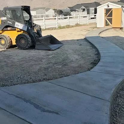 A skid-steer loader working on a curved concrete sidewalk in a backyard, with houses and a shed in the background.