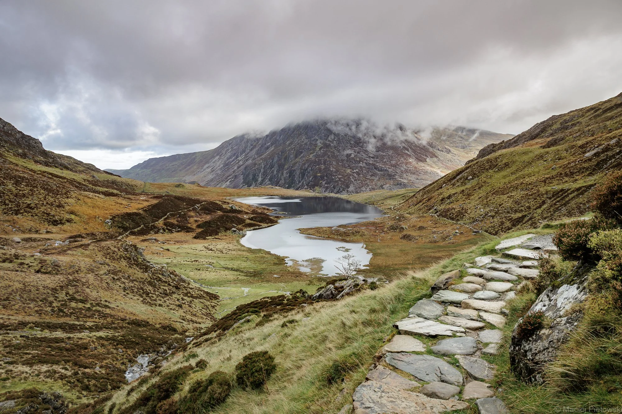 A mountainous landscape with a cloudy sky, a lake in the valley, grassy slopes, and a stone-paved trail along the hillside.