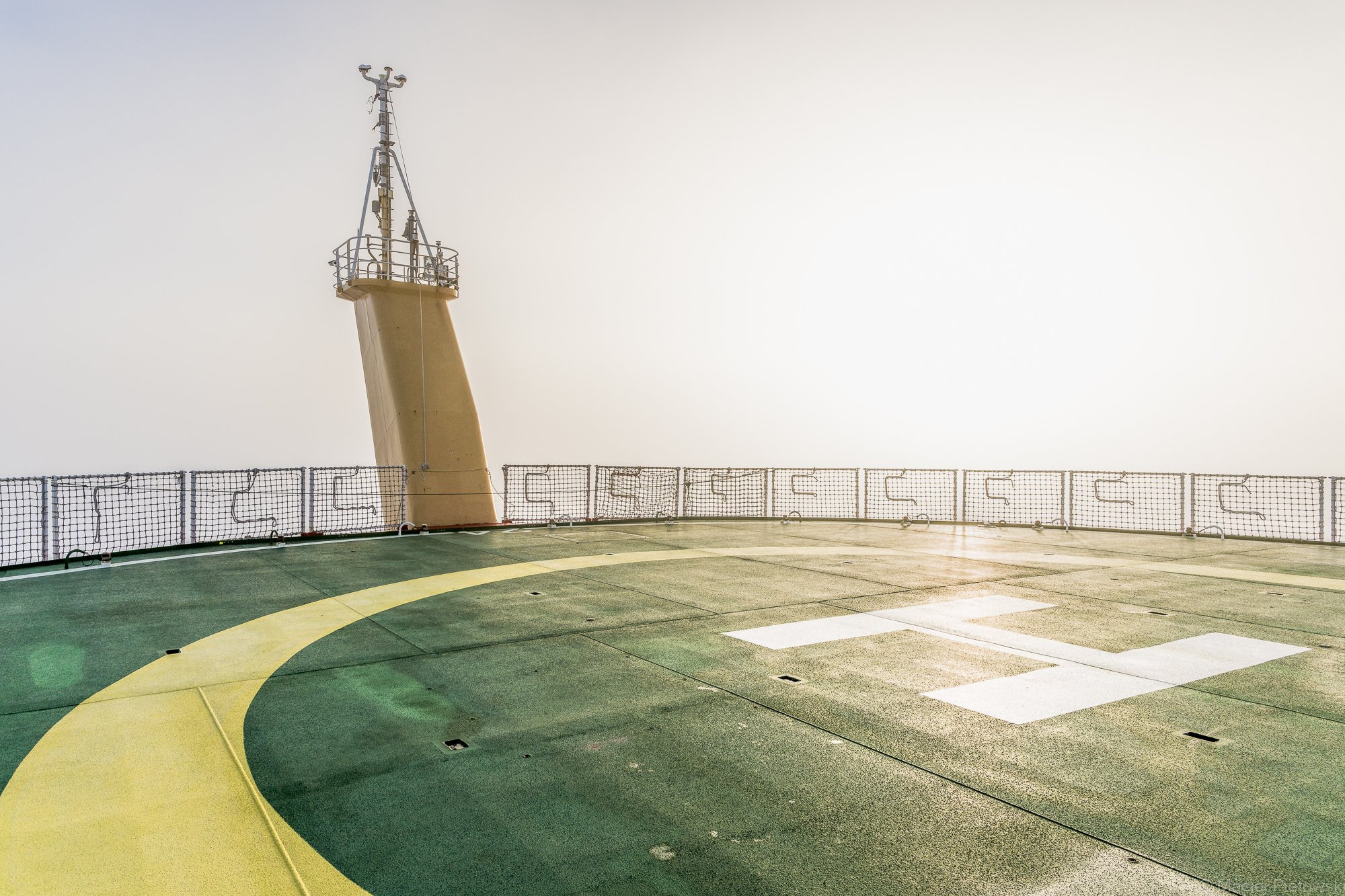 View from the deck of a cruise ship showing a navigation tower and a safety net along the side of the ship. The sky is overcast and the deck surface features painted yellow and green lines.