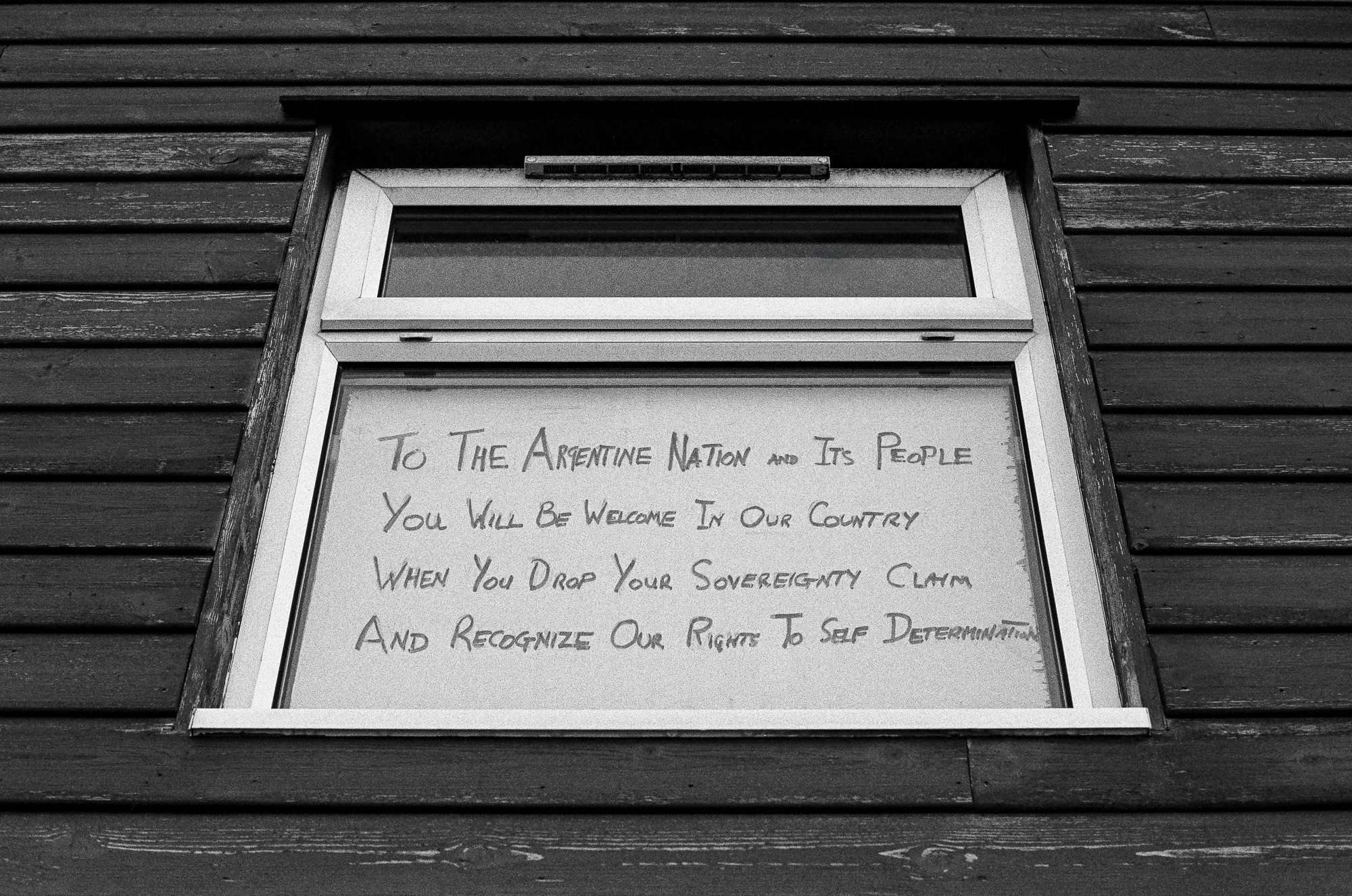 A framed handwritten note on a wall with horizontal wooden planks background. The note begins with 'To the Argentine Nation and Its People...' and mentions welcoming in the country, sovereignty, and self-determination.
