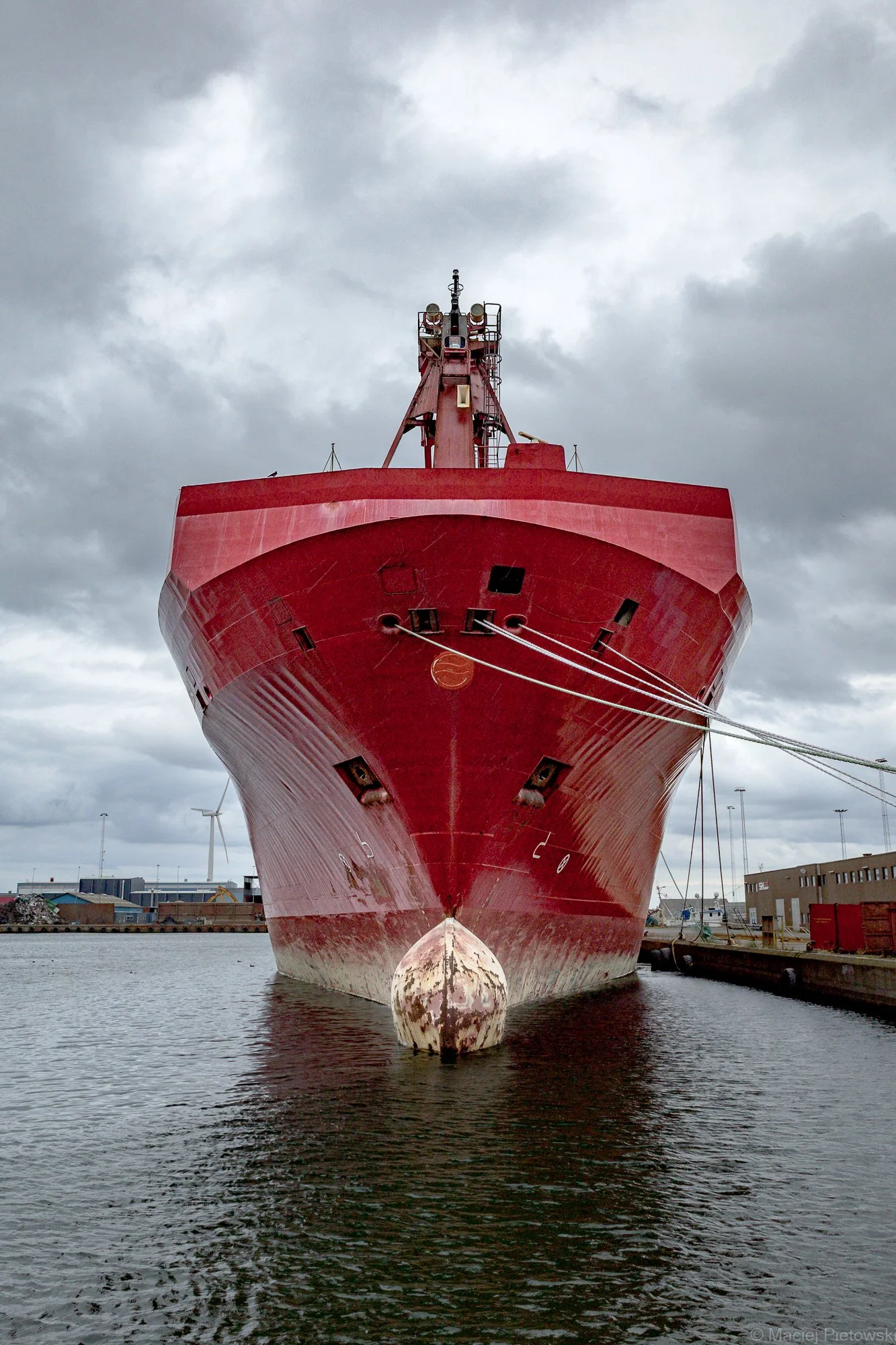 Front view of a large red ship docked at a harbor with cloudy sky overhead.