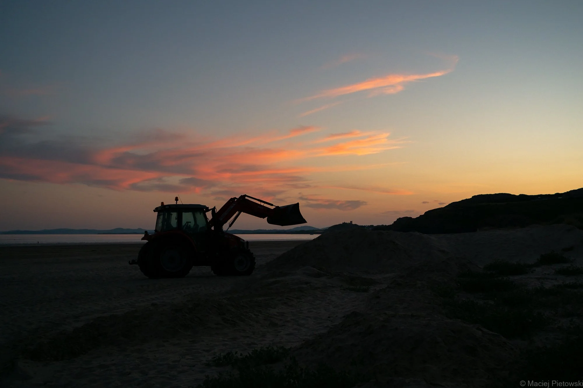 A silhouette of a tractor on a sandy beach during sunset with colorful pink and orange clouds in the sky and distant hills.