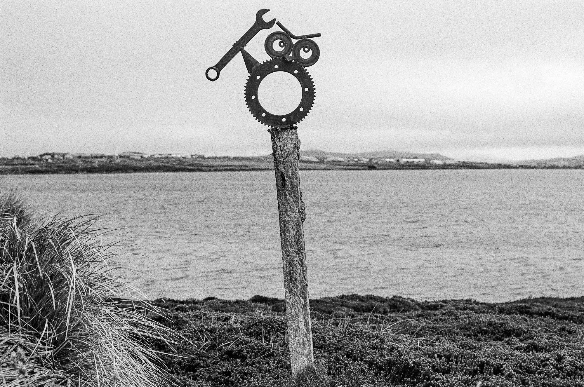 A black and white photo of a decorative sculpture made from bike parts on a wooden post near a body of water with a mountainous landscape in the background.