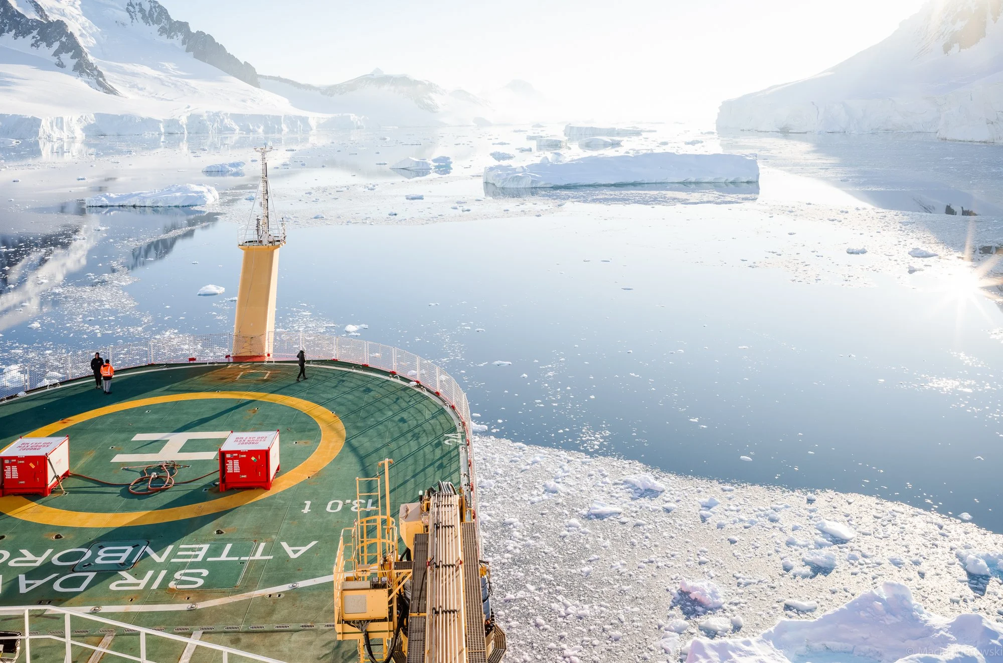 View from a ship's helicopter deck overlooking icy waters with floating icebergs and snow-covered mountains in the background, with sunlight creating a bright atmosphere.
