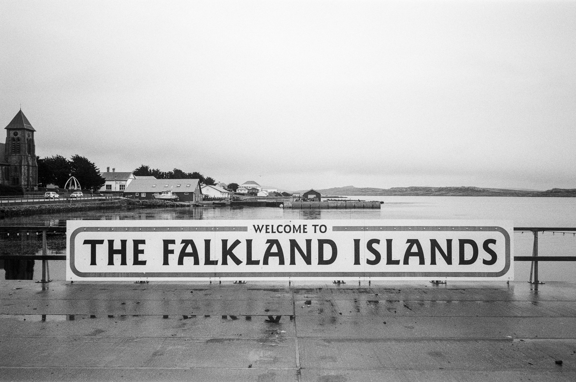 Black and white photograph of a waterfront scene with buildings, a church, and hills in the background. A large sign in the foreground reads "Welcome to the Falkland Islands."
