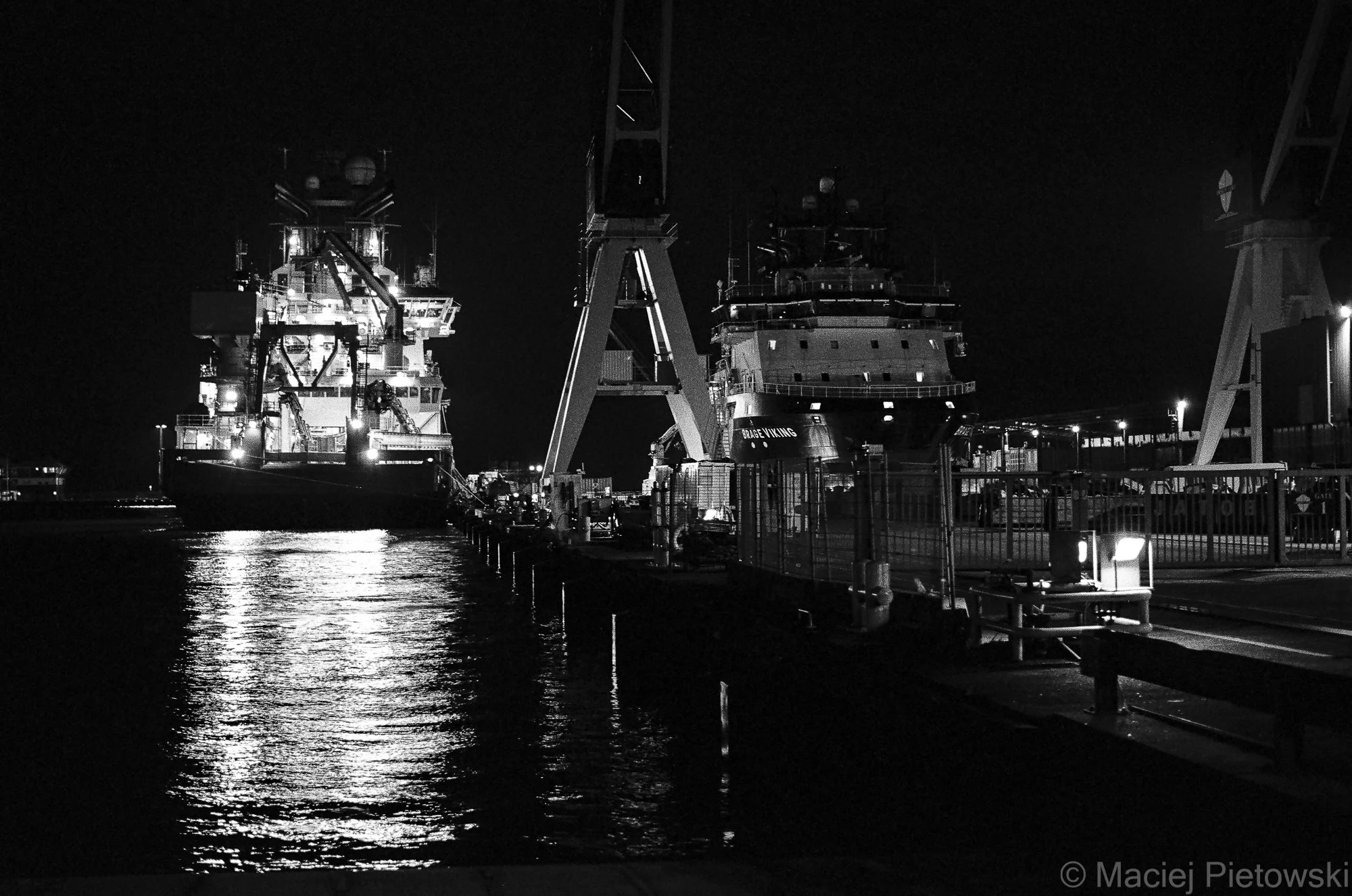 Nighttime view of a dock with large ships and cranes illuminated by artificial lights, water in the foreground reflecting the lights.