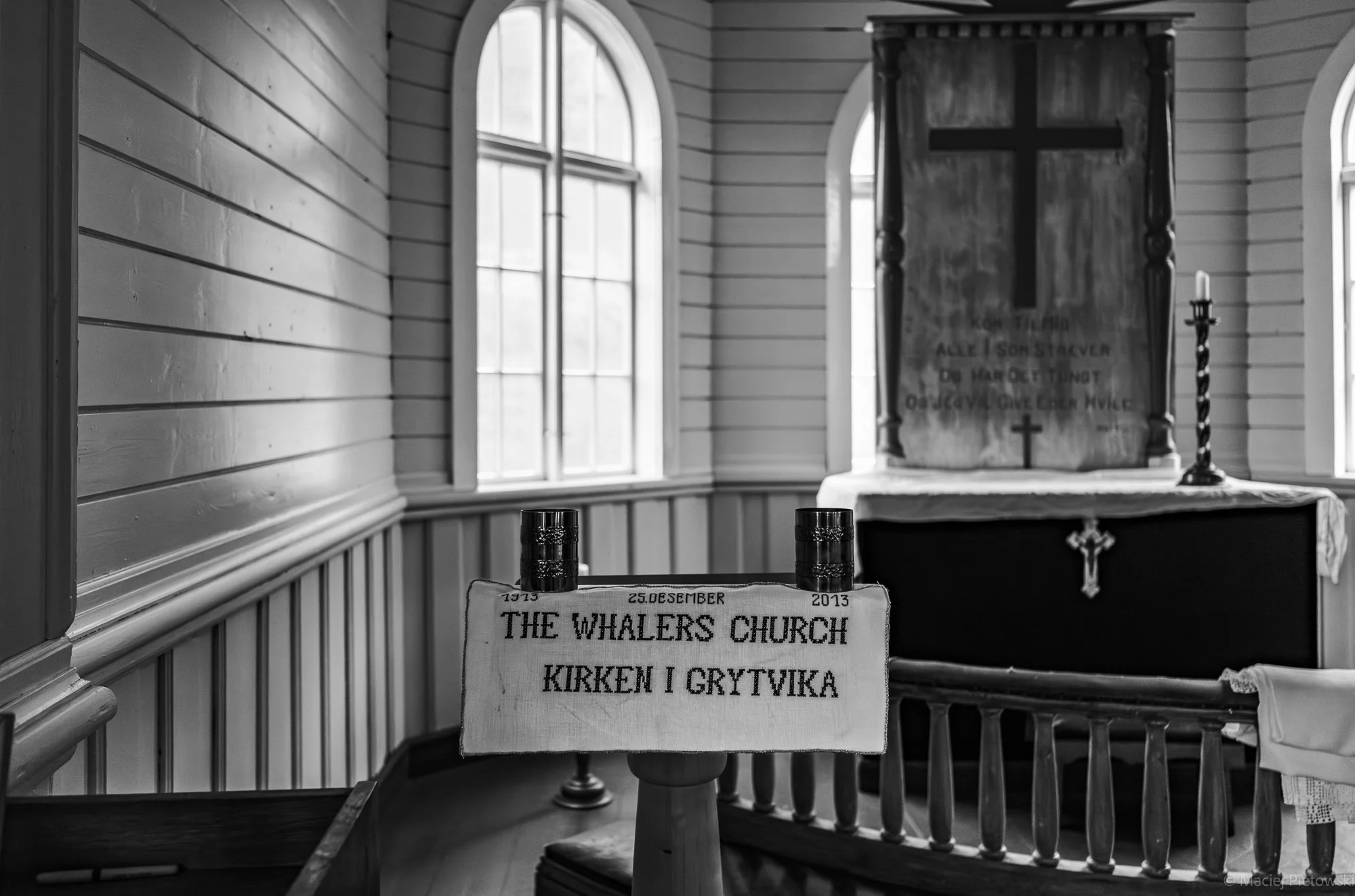Black and white photo of a church interior with arched windows, a wooden cross on a banner, and a table with a cloth displaying the text 'The Whalers Church' and the date 1913, December 25, 2013.
