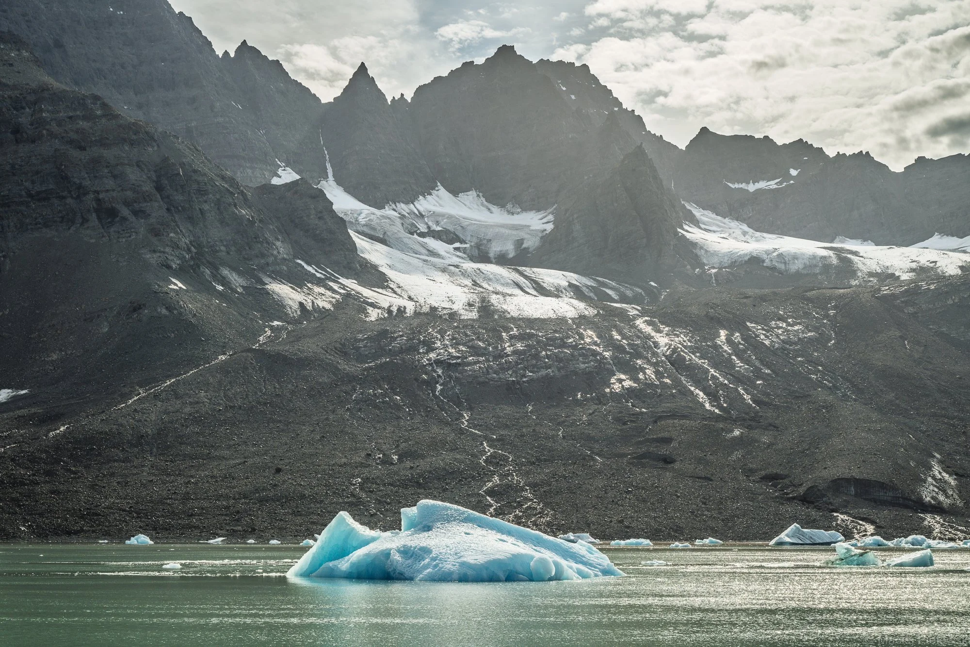 A glacier meeting a body of water with icebergs floating, mountains in the background, and snow patches on the rocky mountain slopes under a cloudy sky.
