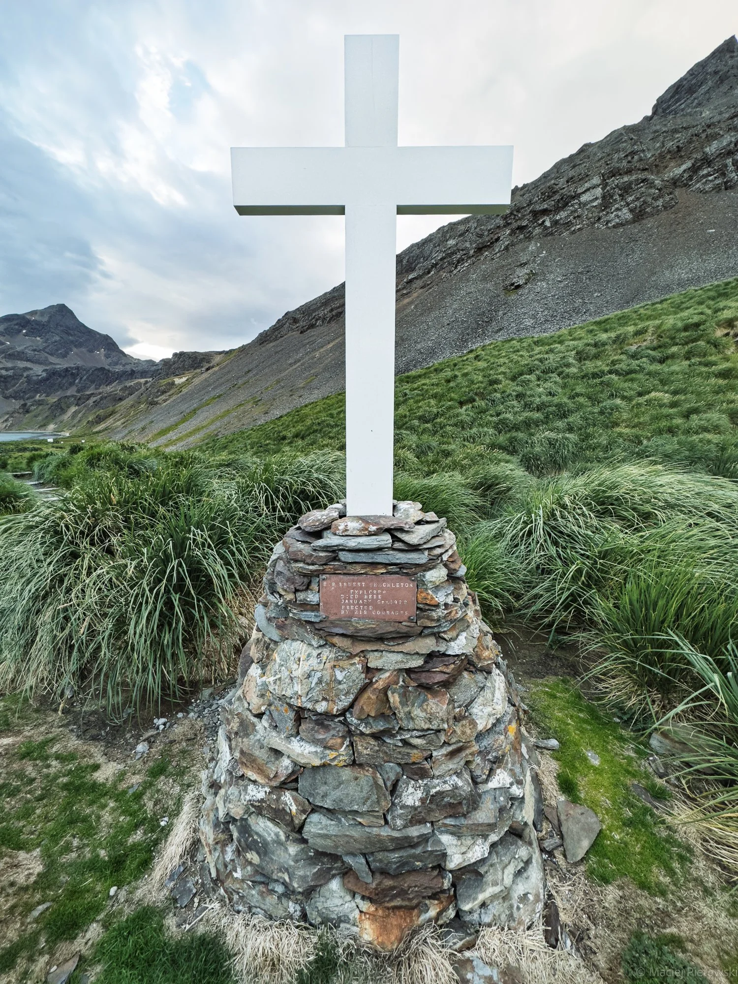 White cross monument on a pile of rocks, with grassy area and mountains in the background.