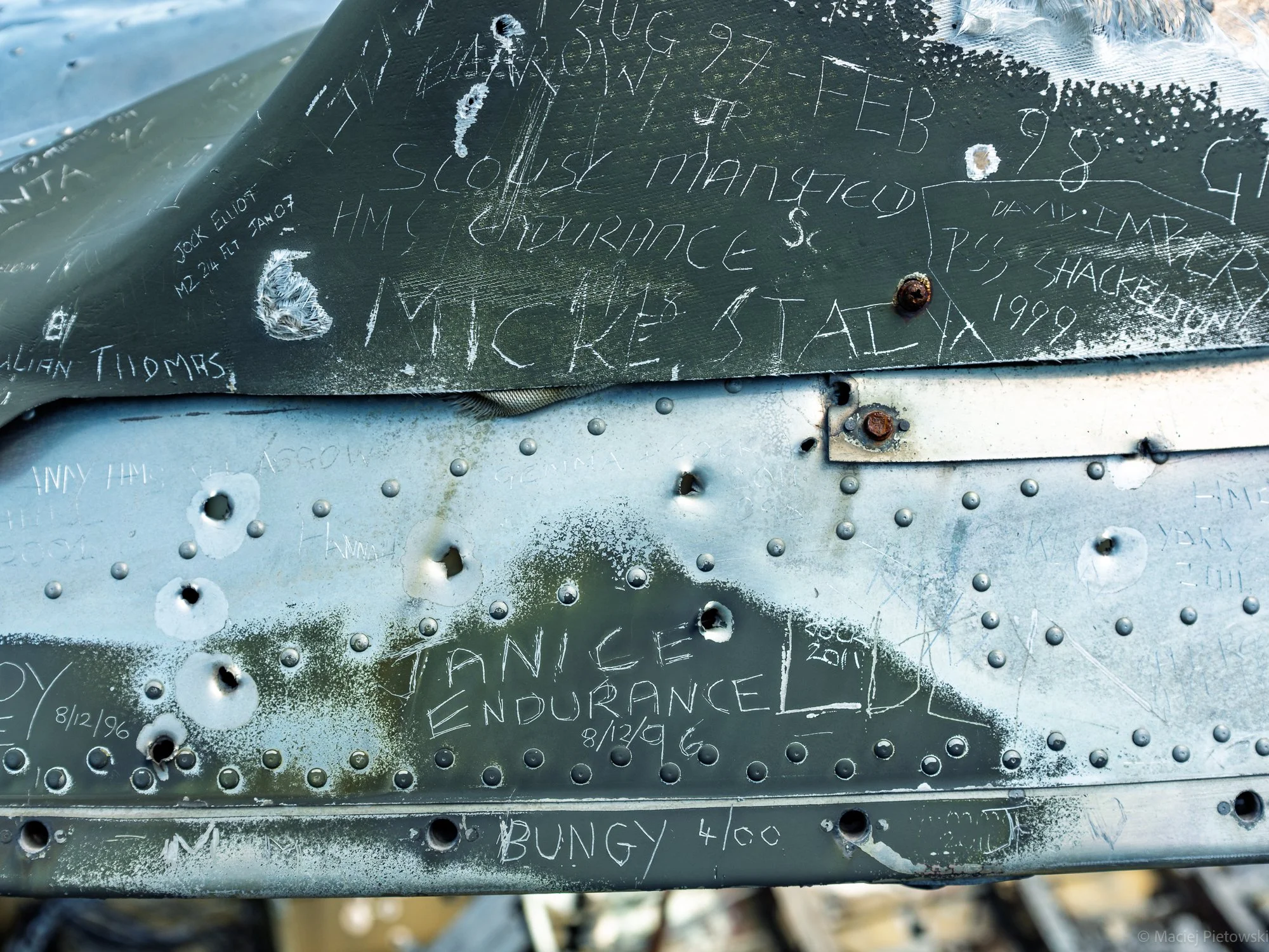 Close-up of a weathered metal surface with numerous handwritten notes and signatures scratched into it. The writing includes dates, names, and locations, some of which are faded or partially obscured by rust and scratches.