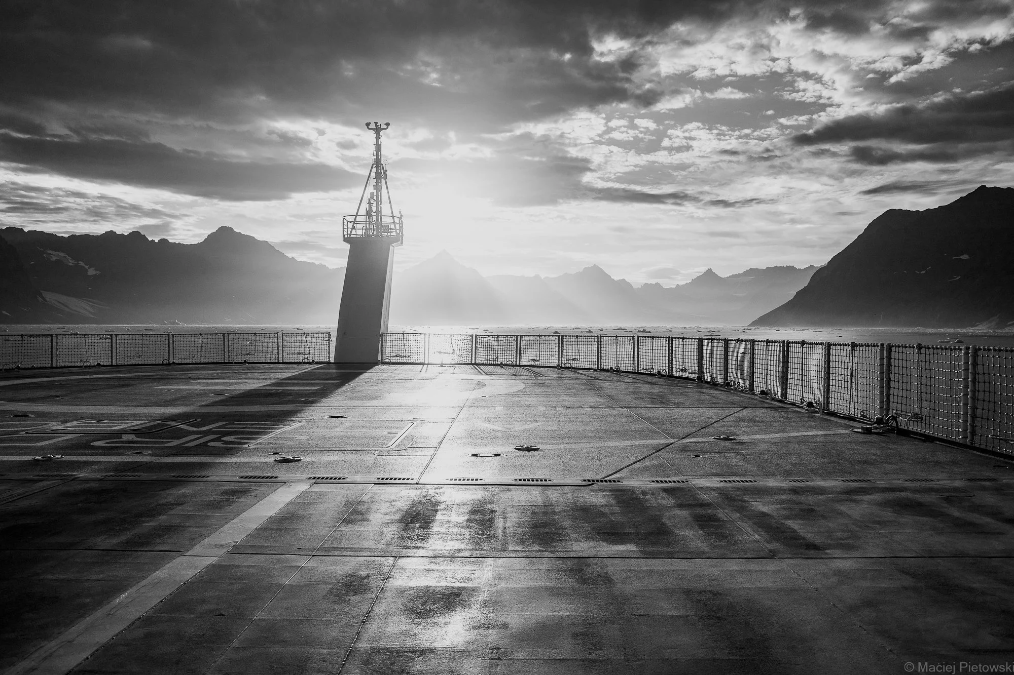 A black-and-white photo of an aircraft carrier deck with a lighthouse structure near the water, surrounded by mountains and cloudy sky in the background.