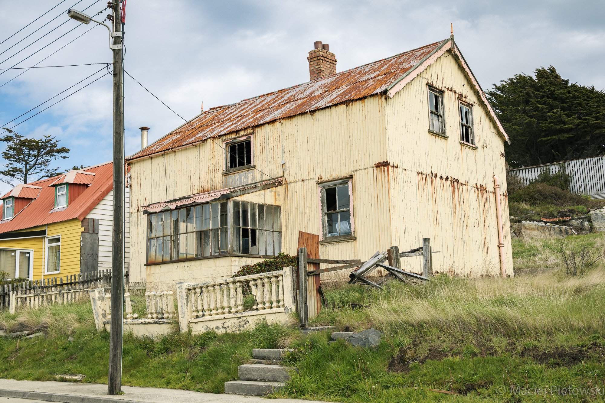 Old, abandoned house with peeling yellow paint, broken windows, and rusted corrugated metal roof. Overgrown grass and a white wooden fence surround the house, with a set of concrete steps leading up to the front entrance, and a tall utility pole with