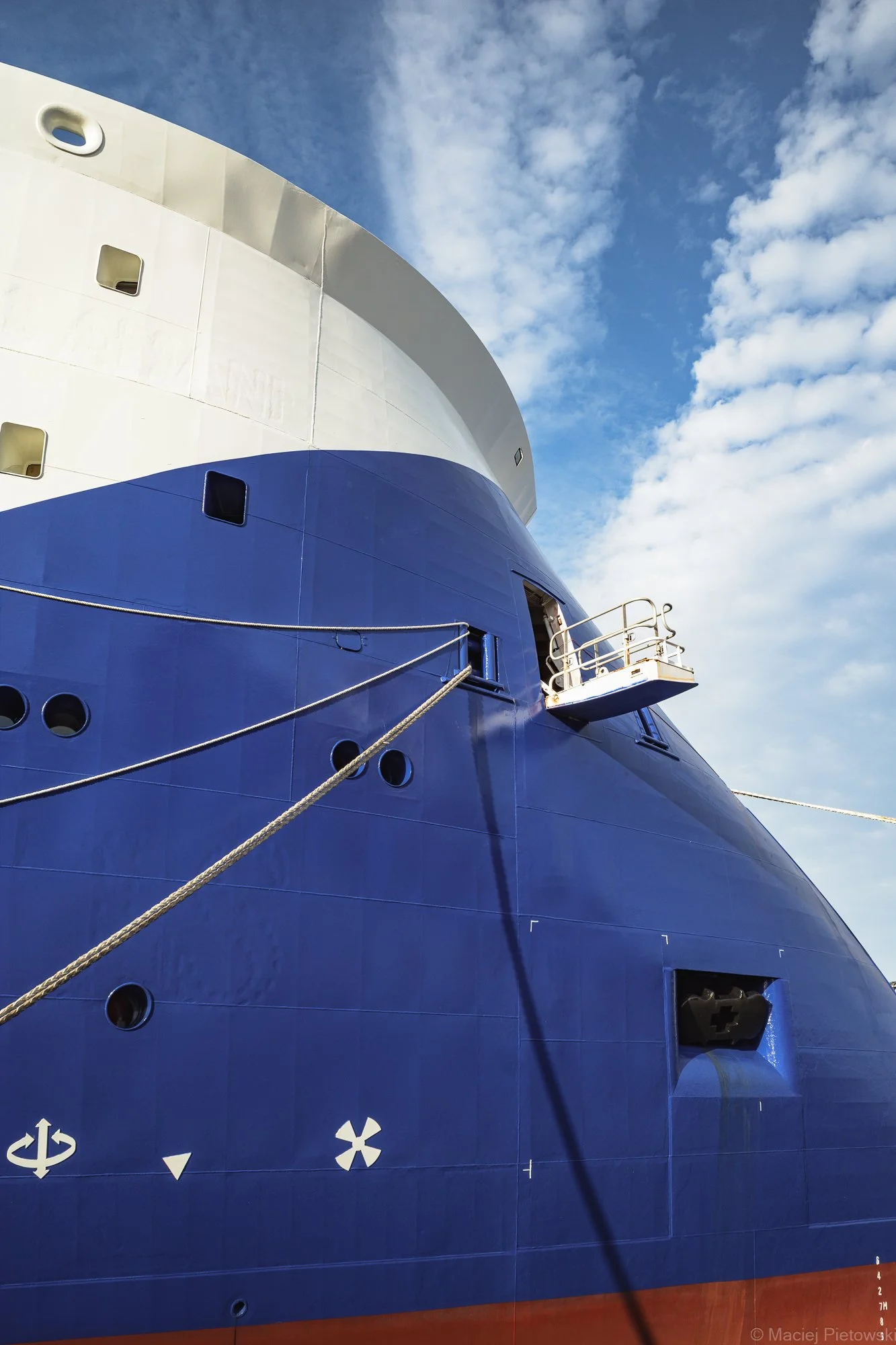 Close-up of a large ships hull painted in blue and white with the waterline marked in red, with various symbols and ropes attached.