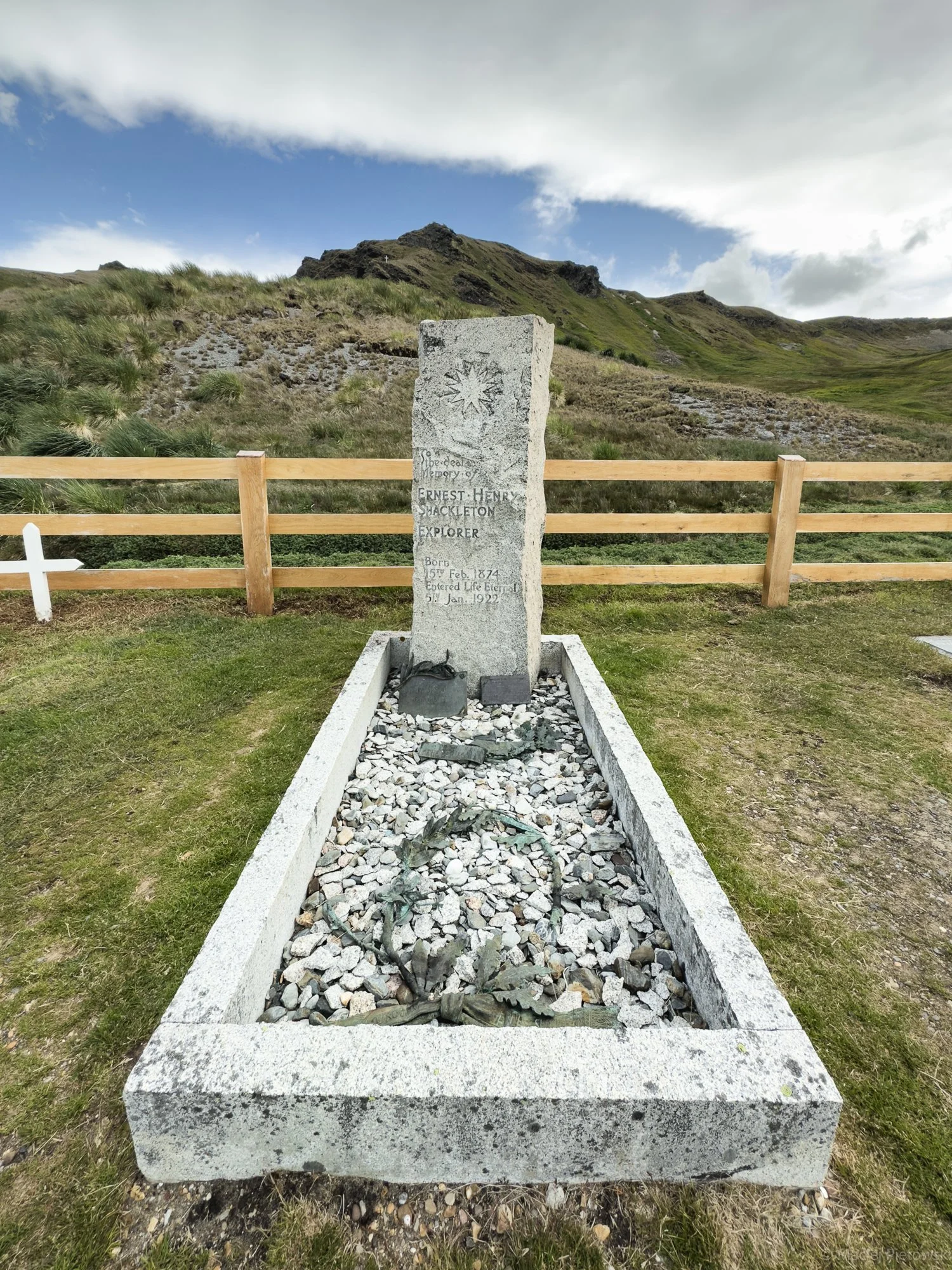 A memorial stone with an engraving dedicated to explorer Ernest Henry Shackleton, located in a grassy area with a mountain in the background and a partly cloudy sky.