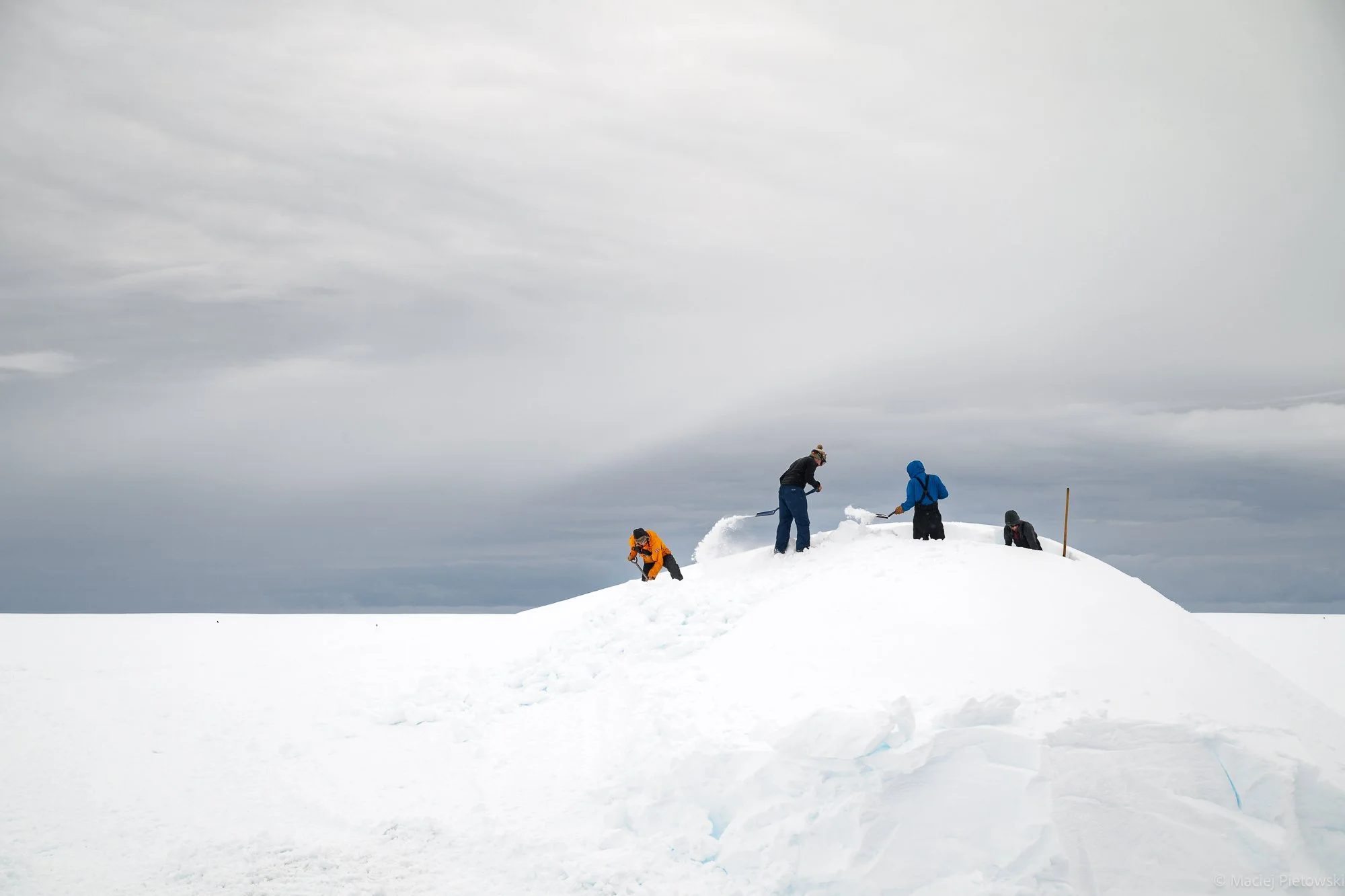 Group of people in winter clothing shoveling and clearing snow on a snow-covered landscape under a cloudy sky.
