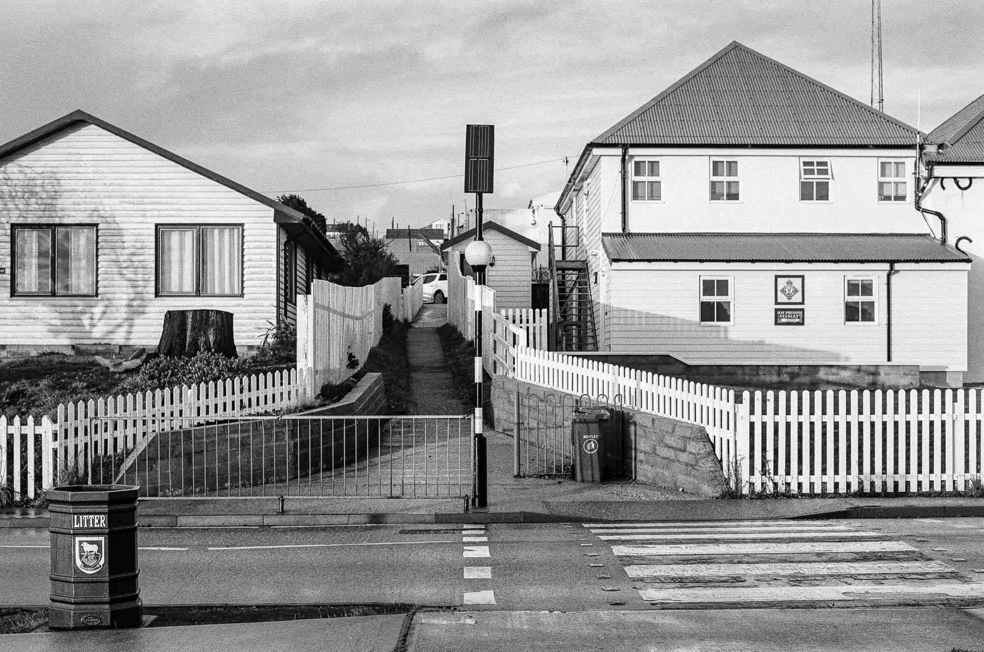 Street scene with a white picket fence, trash and recycling bins, houses, and a pedestrian walkway. There is a street crossing with a pedestrian crossing signal in the foreground and a large building with multiple windows in the background.