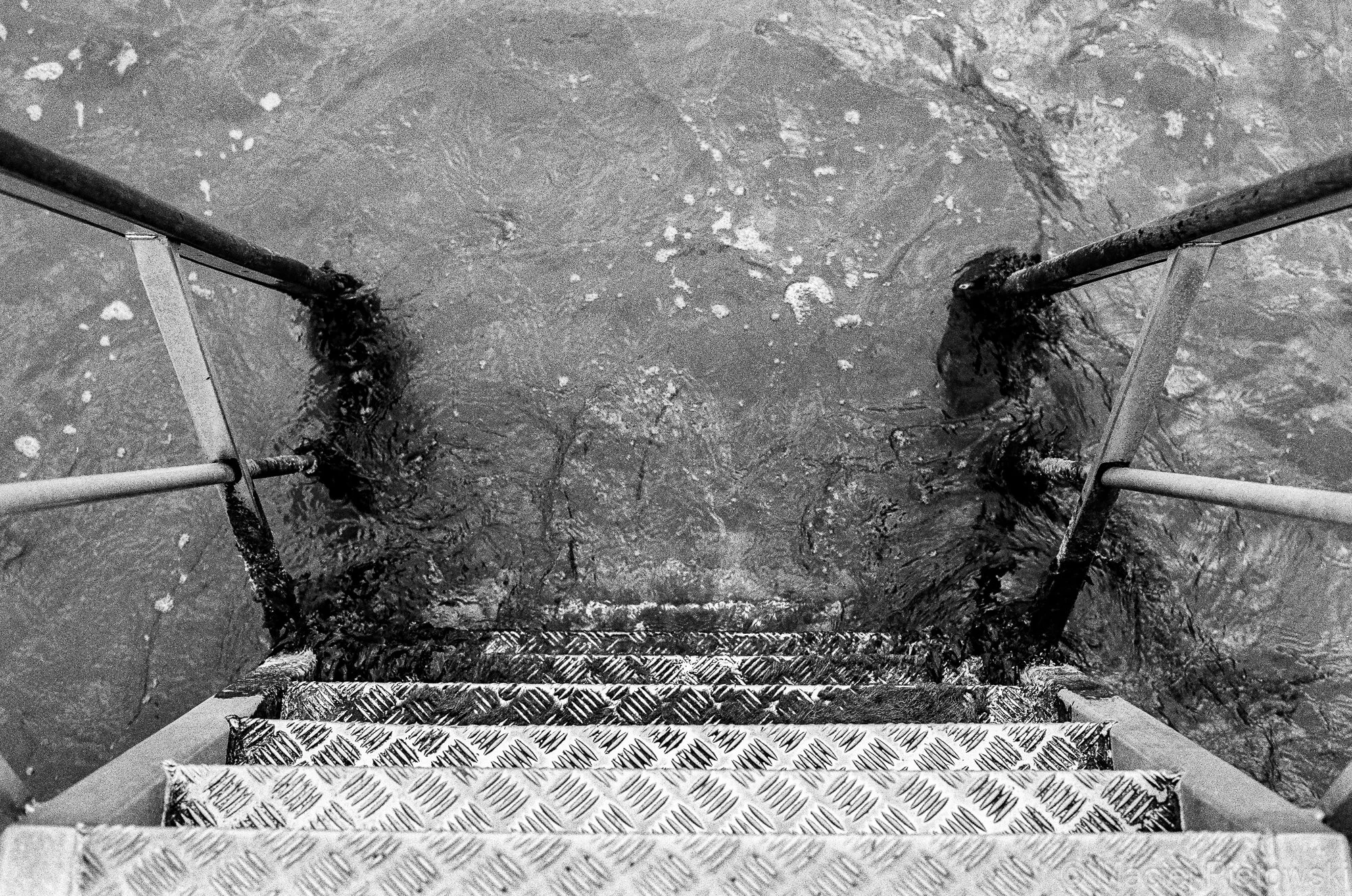 Black and white photo taken from the top of a metal staircase looking down at a body of water. The staircase has a textured, diamond-plate surface and metal railing, and appears wet, with water collected on the steps. The water below is rippled, show