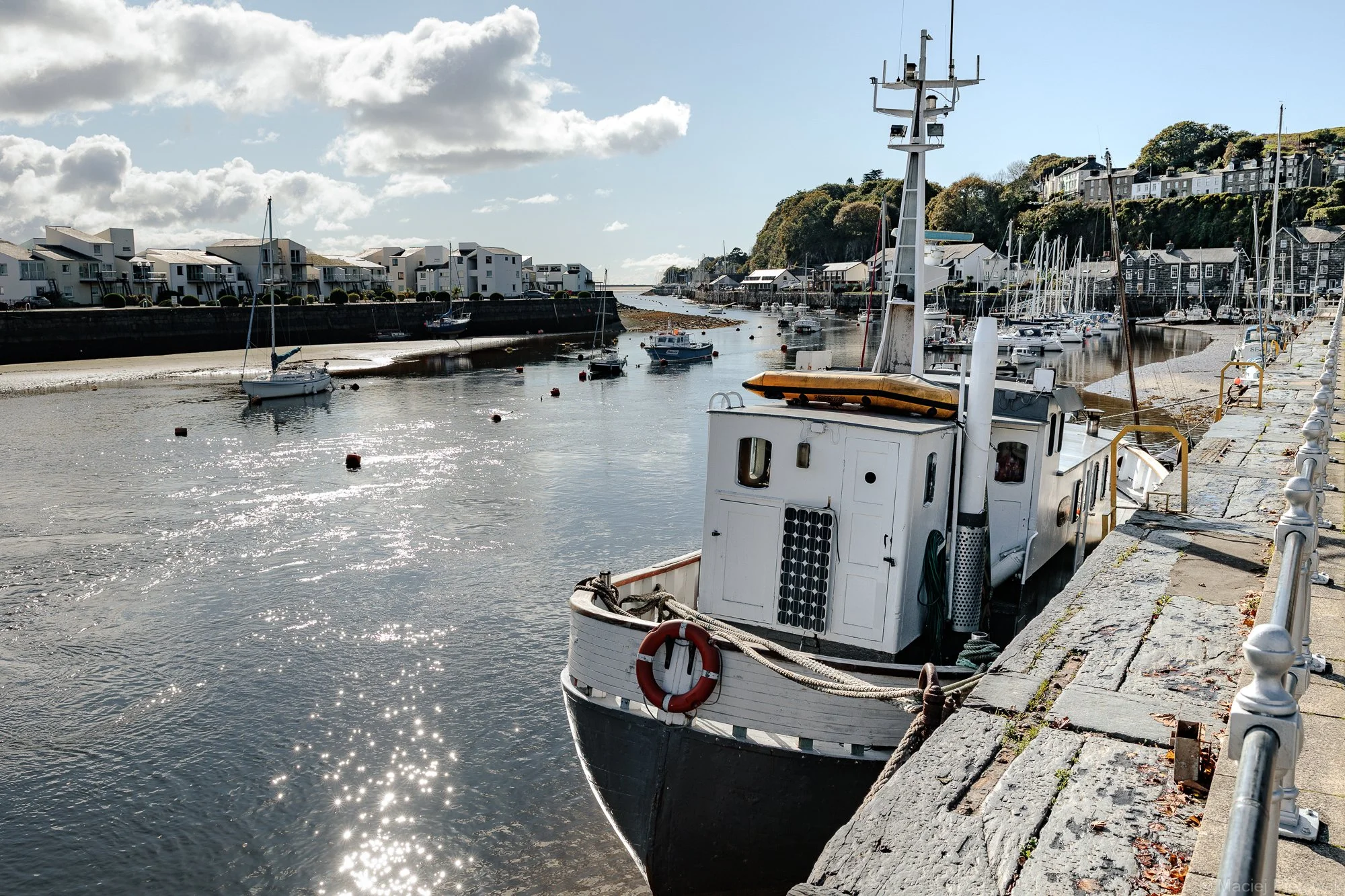 A small harbor with boats docked along the pier, residential buildings on a hillside, and a partly cloudy sky reflecting sunlight on the water.
