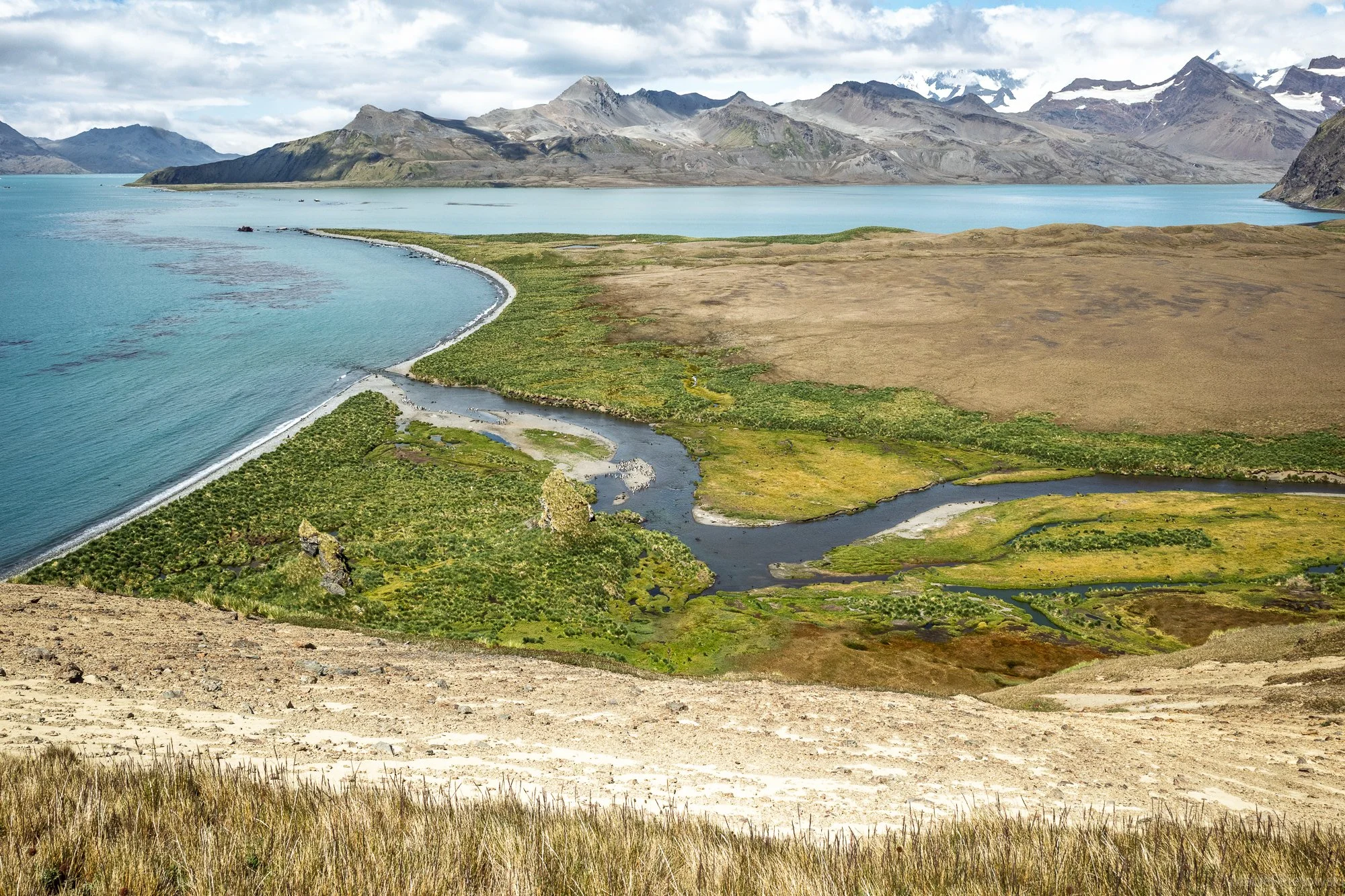 Panoramic view of a mountainous landscape with a large body of water, green and brown fields, and a meandering river in the foreground. The sky is cloudy and snow-capped mountains are visible in the background.