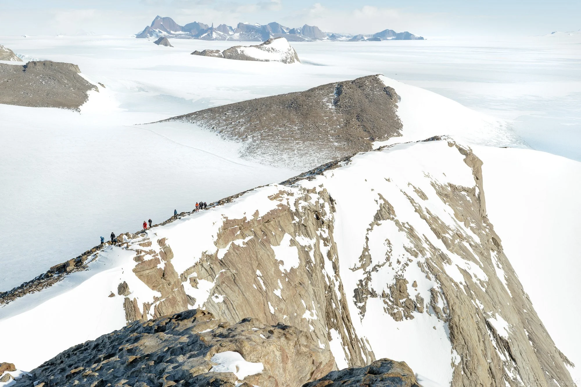 A group of hikers traversing a snow-covered mountainous ridge with steep rock faces, with snowy terrain and icy landscape stretching into the distance under a pale sky.