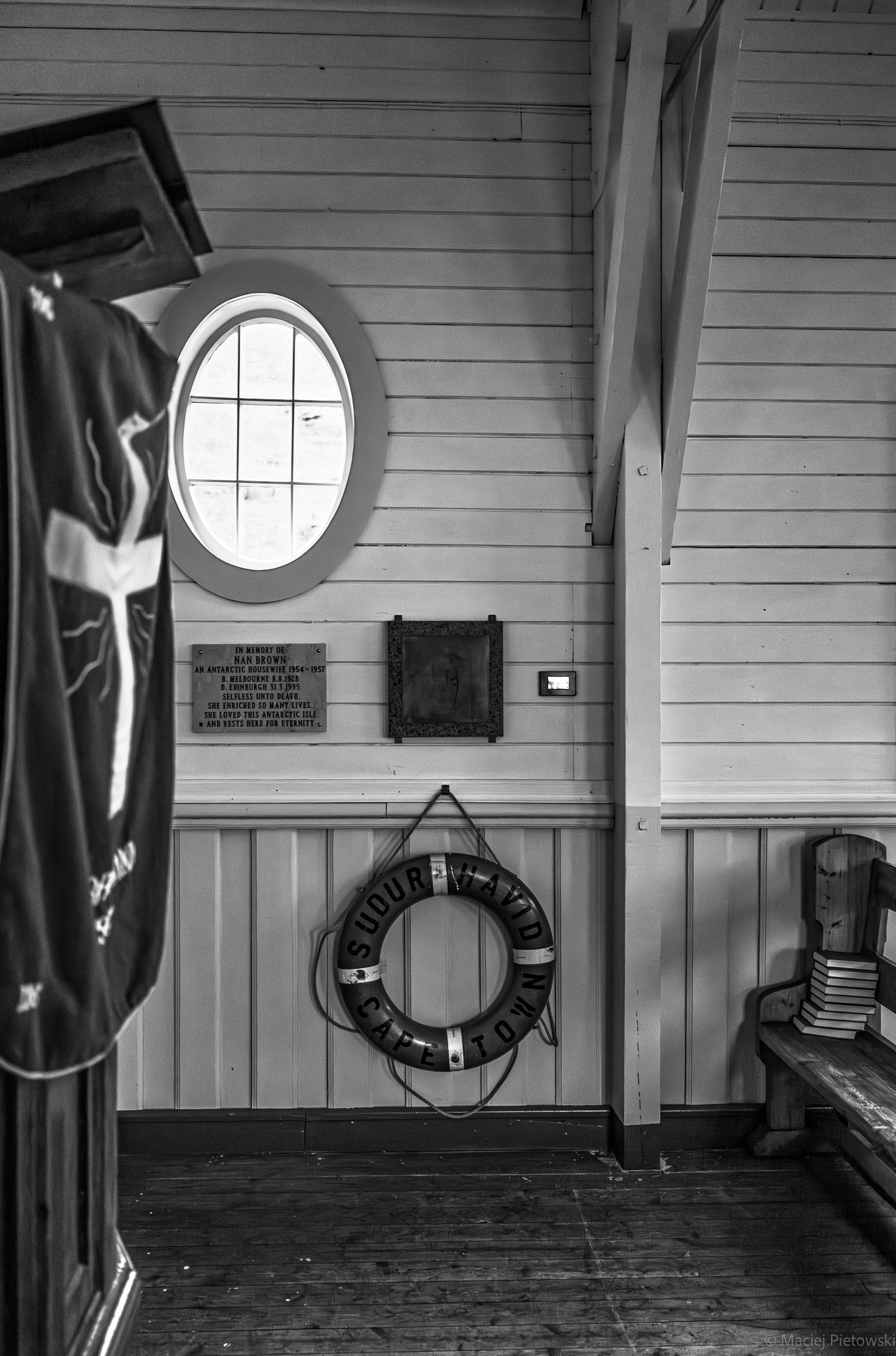 Interior of a wooden building with a circular window, framed by horizontal wood panels; a plaque with memorial text on the wall; a lifebuoy with the text 'SOUR HAVID CAPE TOWN' hangs on the wall; part of a chair and a bookshelf are visible at the rig