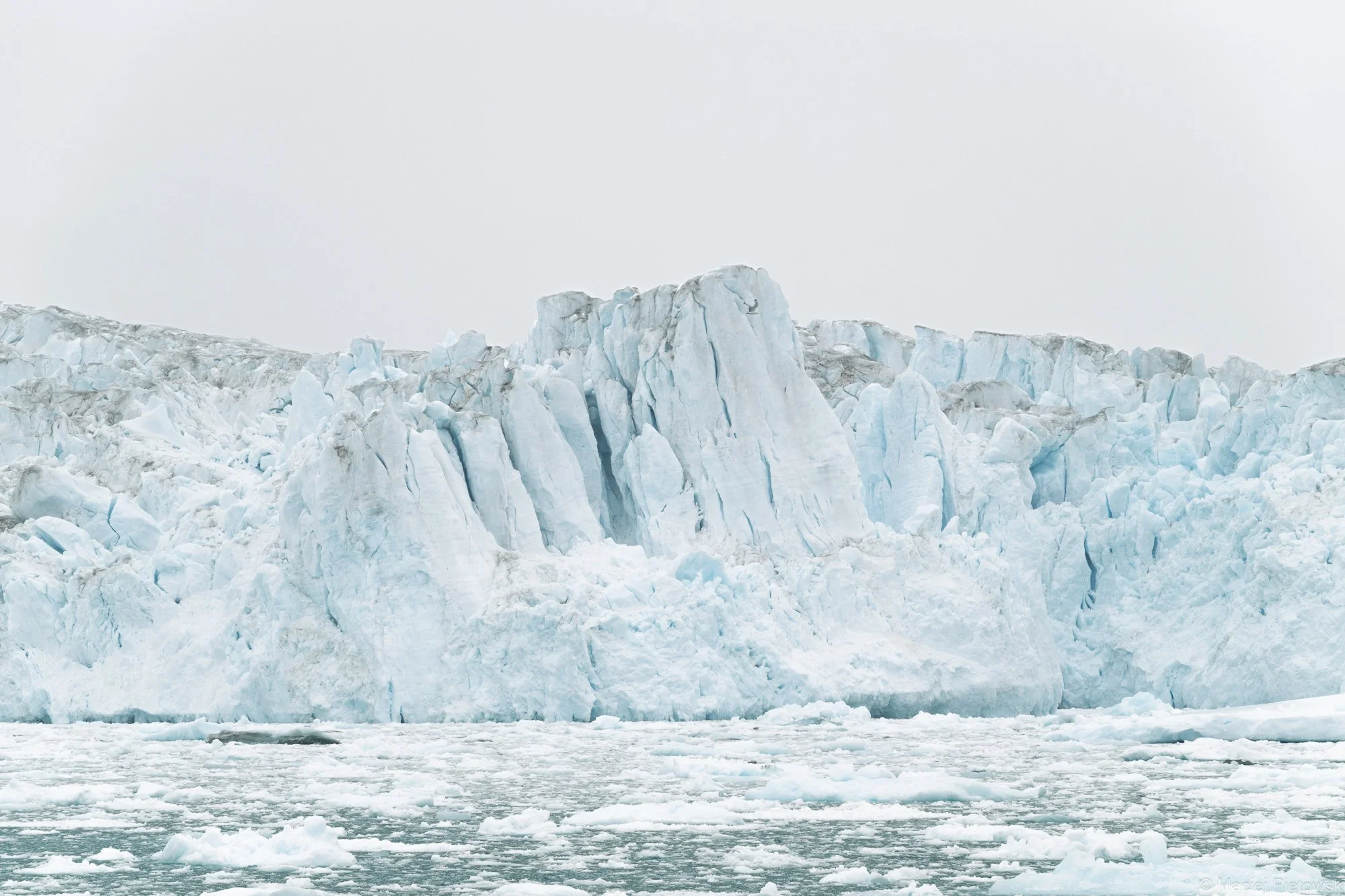 A mountain of glacier ice with jagged and crevassed blue and white ice formations, with a body of water and floating ice pieces in the foreground under a cloudy sky.