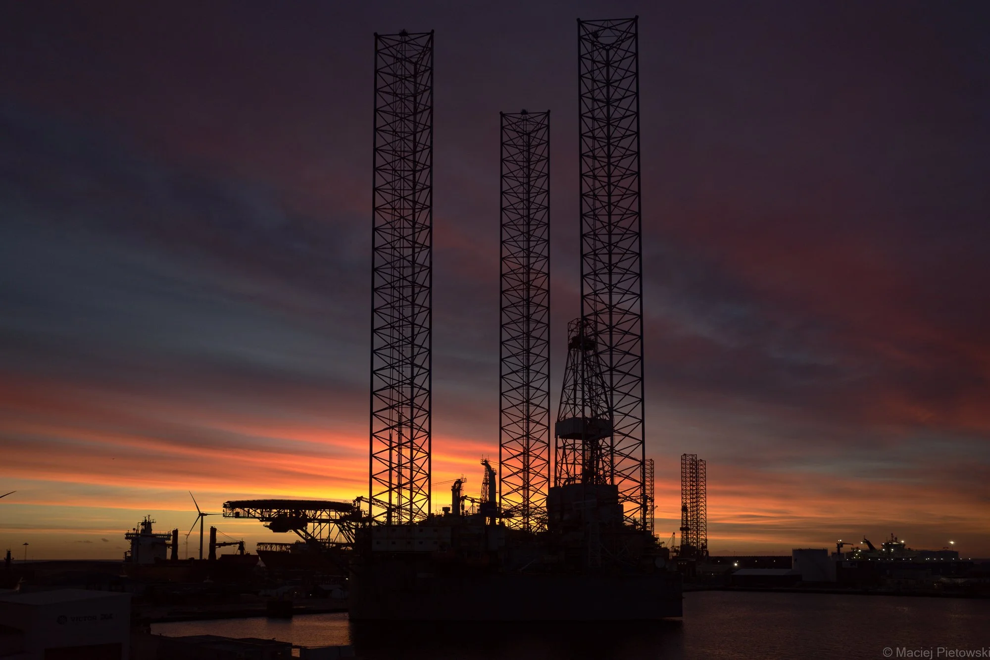 Sunset over an offshore oil platform and telecommunications towers with a wind turbine in the distance.