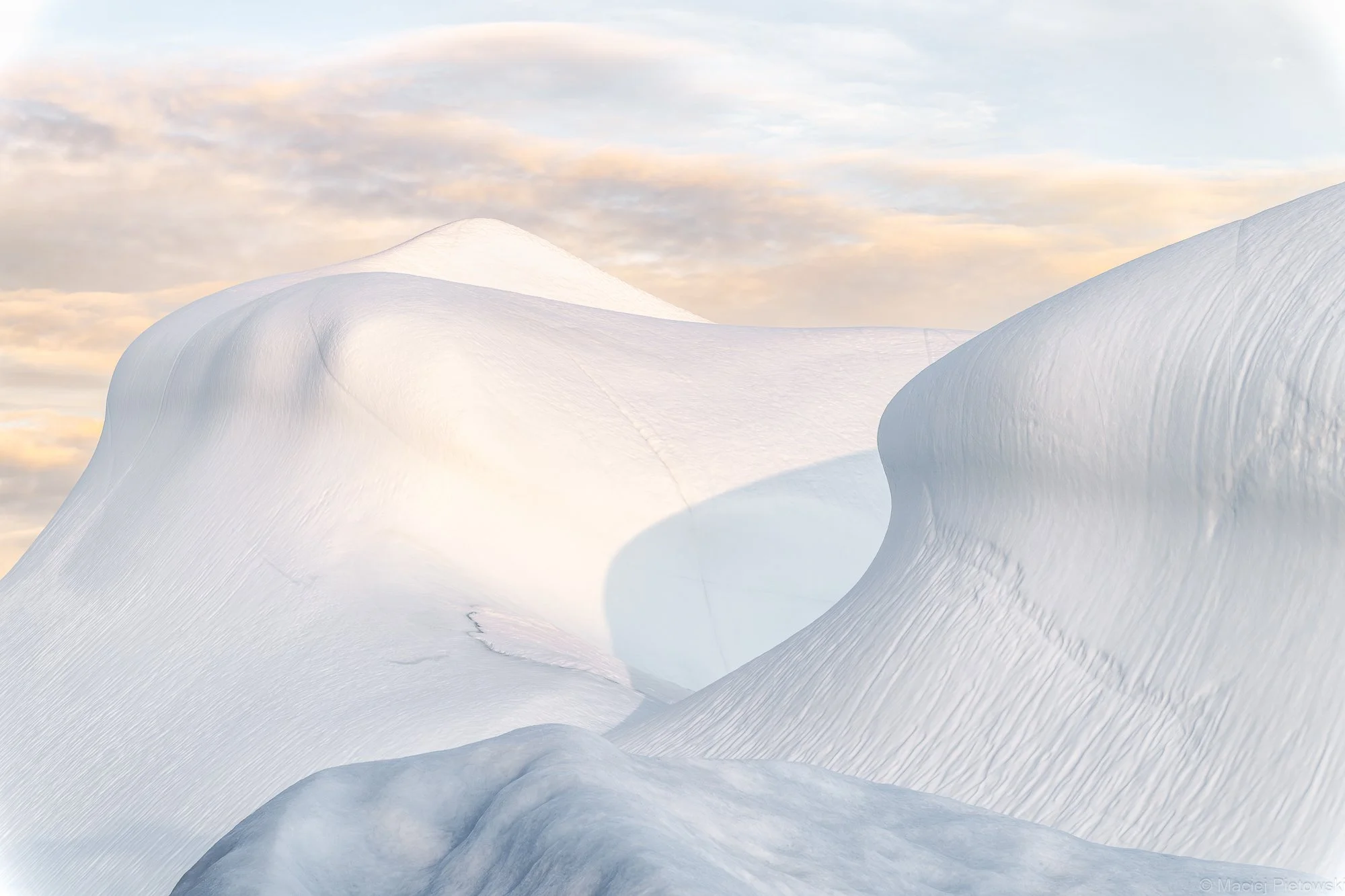 Snowy mountain landscape with smooth, curved snow formations under a partly cloudy sky during sunset.