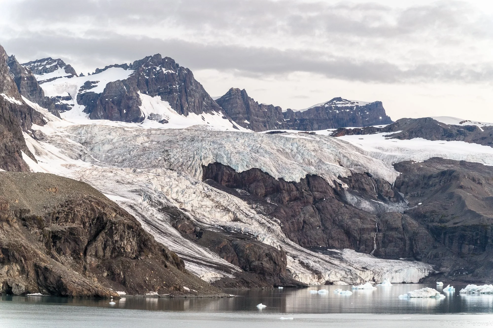 Glacier calving into a body of water in a mountainous landscape under a cloudy sky.